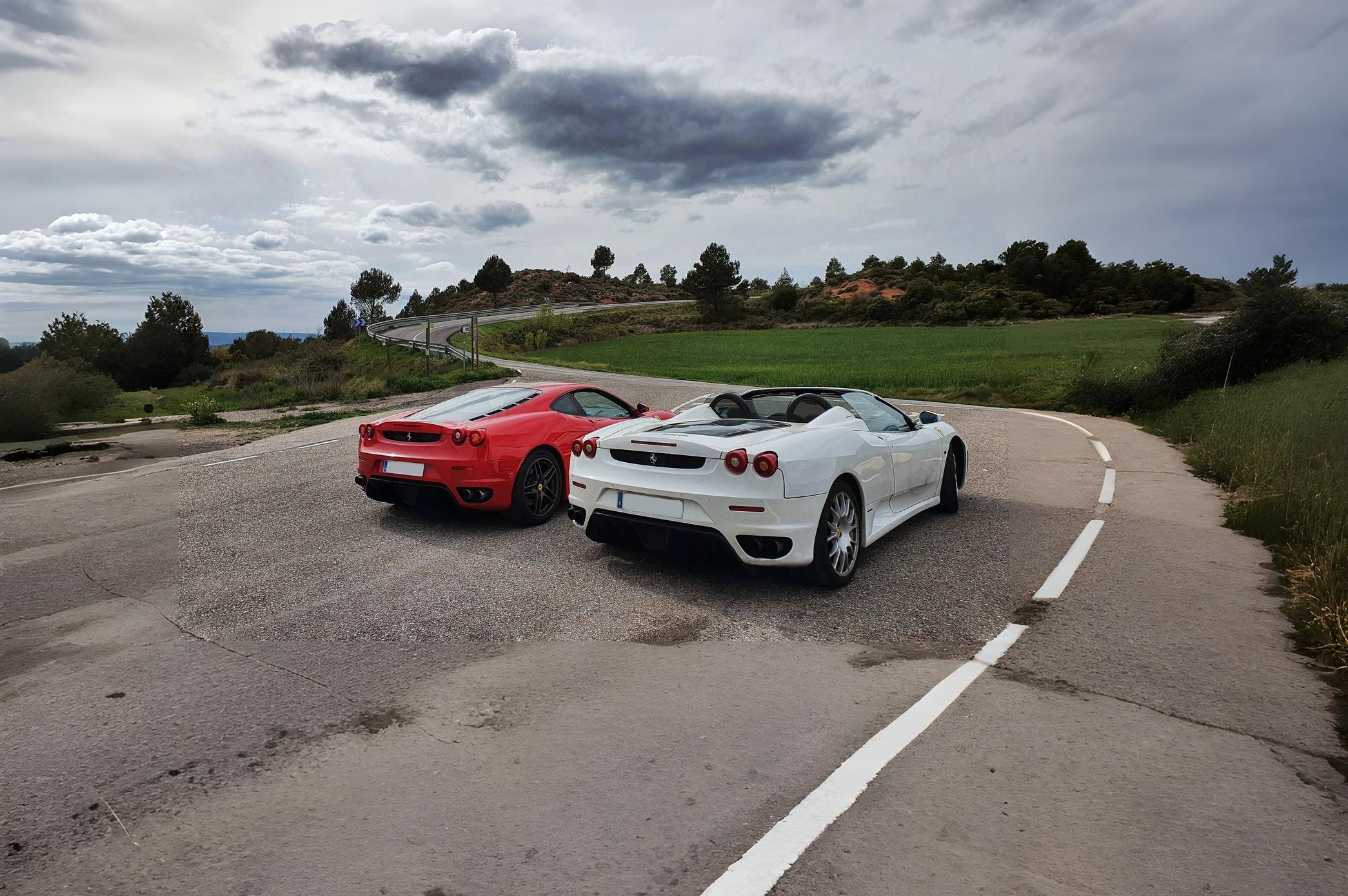 Dos coches deportivos, uno rojo y otro blanco, estacionados en una carretera rural con paisaje campestre y cielo nublado de fondo.
