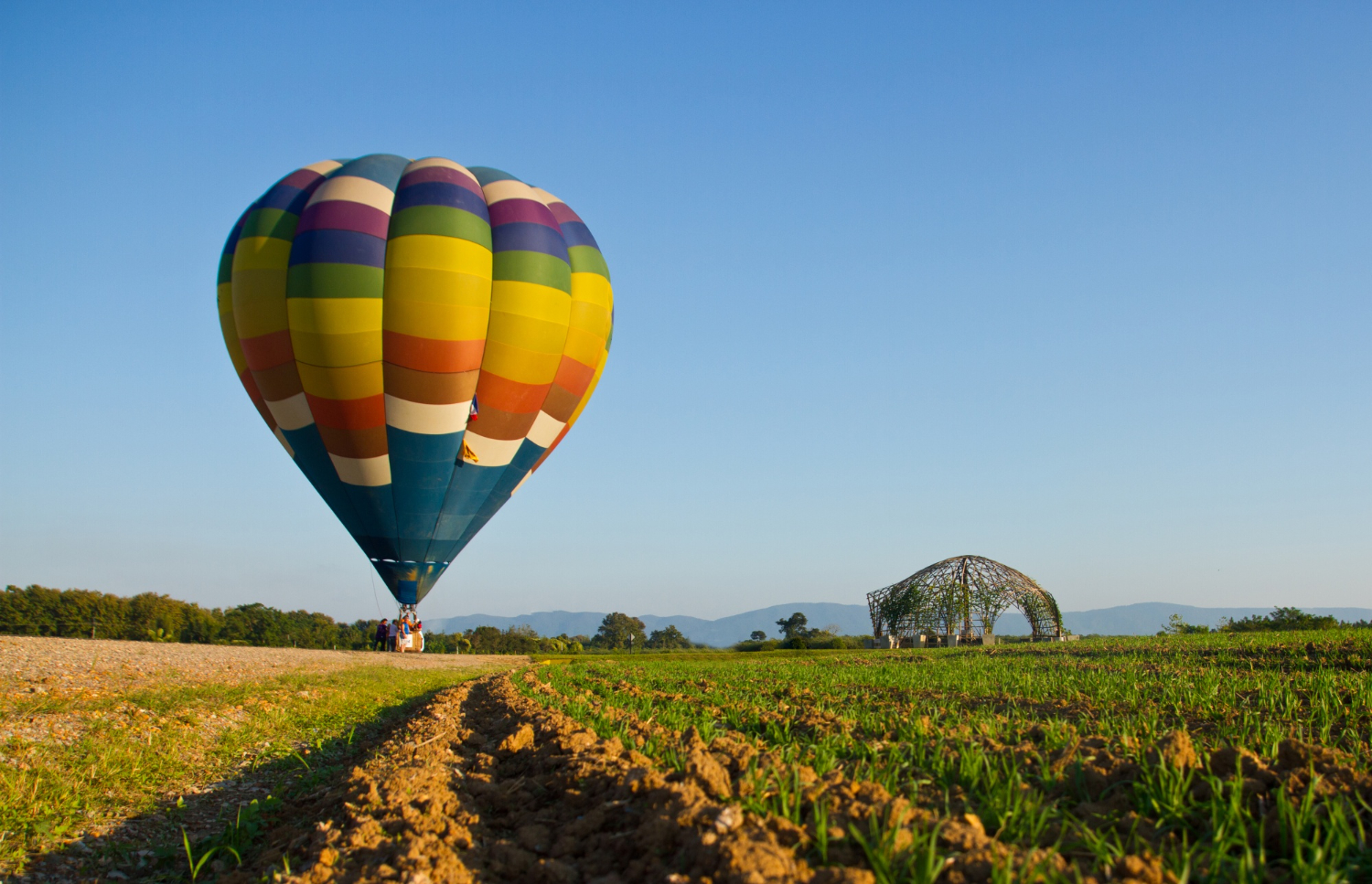Globo aerostático multicolor listo para despegar sobre un campo verde, con una estructura artística al fondo y cielo despejado