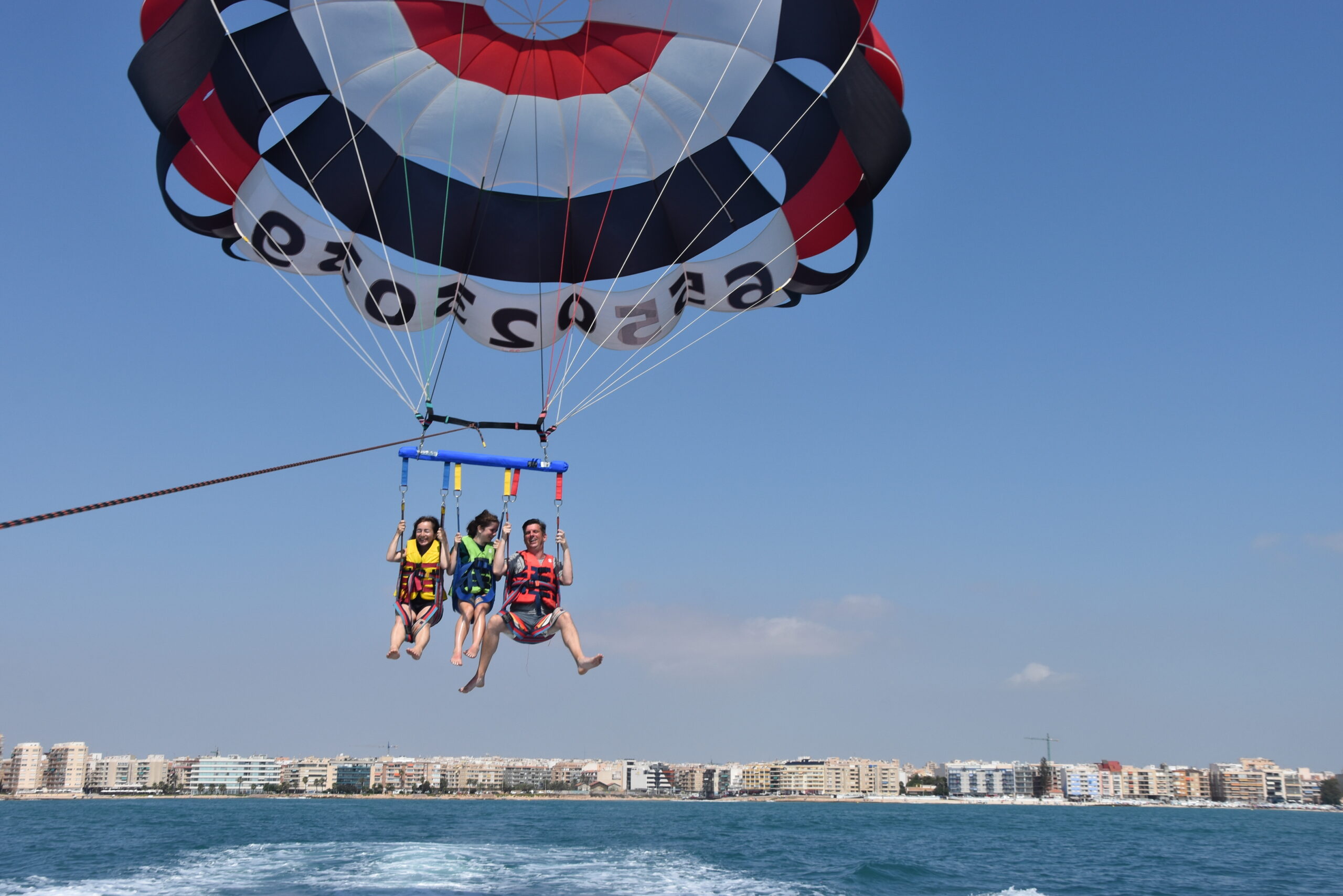 Tres personas disfrutan de una experiencia de paracaidismo ascensional, suspendidas en el aire sobre el agua bajo un cielo azul despejado, con la costa visible a lo lejos.