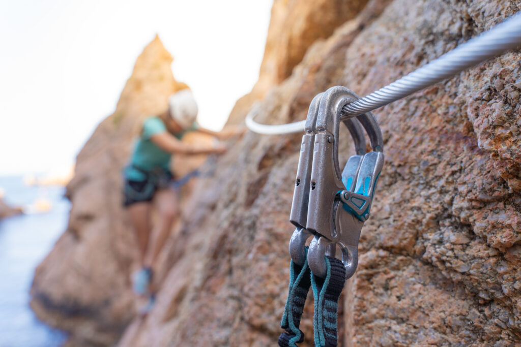 Mujer realizando una vía ferrata junto al mar, equipada con casco y cuerda, sobre rocas al sol