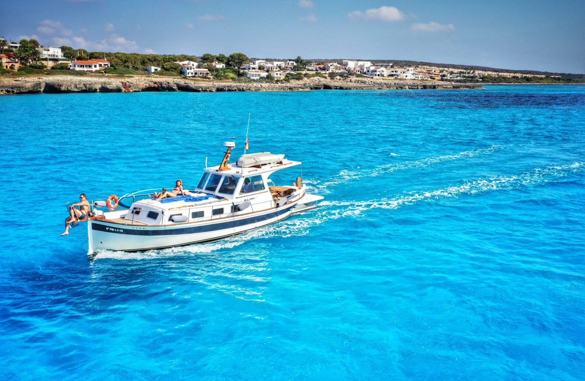 Barco de recreo con personas tomando el sol navegando por aguas turquesas cerca de la costa mediterránea con casas blancas al fondo.