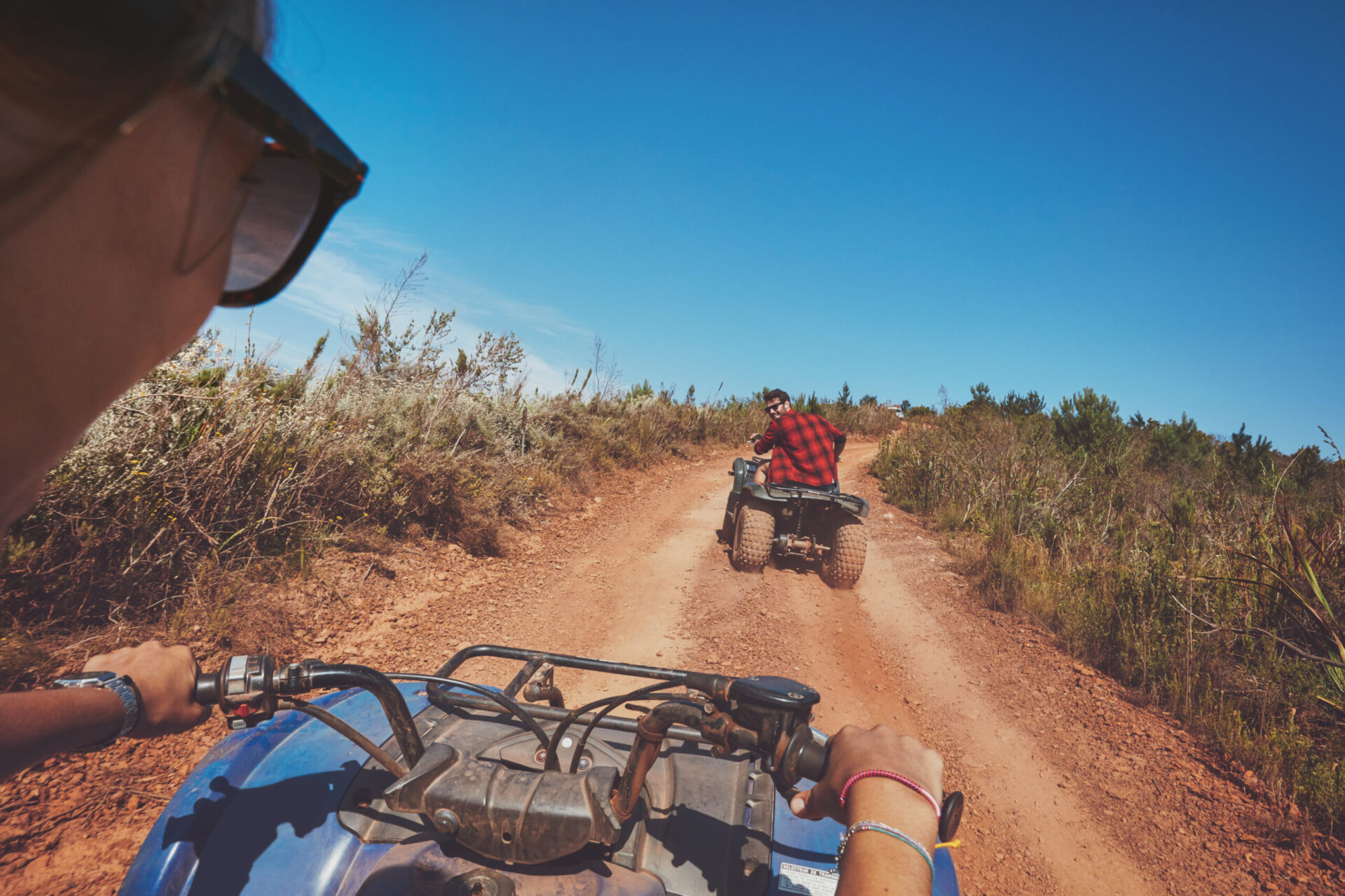 Dos personas conduciendo quads por un camino de tierra rodeado de vegetación y montañas al fondo
