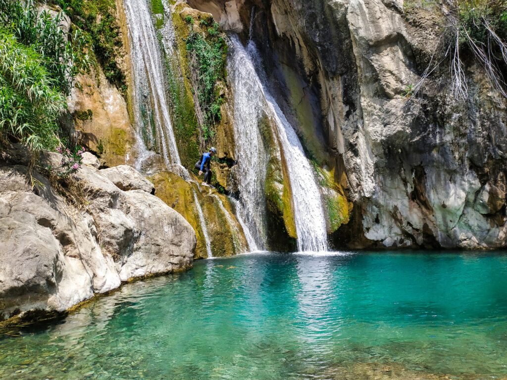 Persona saltando a una poza de agua turquesa mientras un grupo con trajes de neopreno observa desde la orilla, en un entorno natural de montaña.