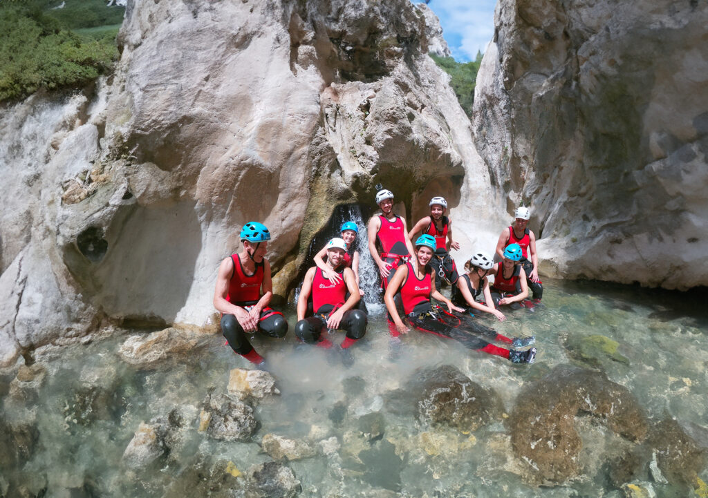 Grupo de personas con trajes de neopreno rojos y cascos, sentados en el agua entre formaciones rocosas durante una ruta de barranquismo.