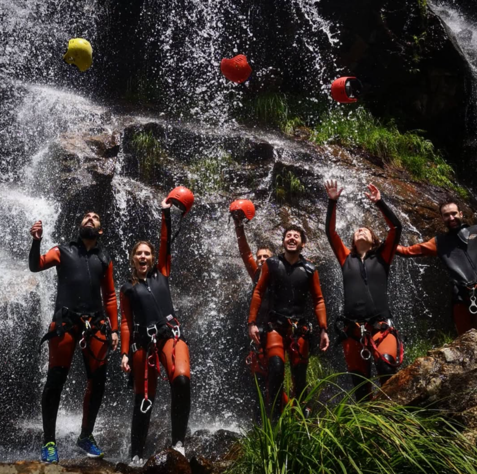 Grupo de personas con trajes de neopreno y cascos naranjas celebrando frente a una cascada, levantando los brazos y lanzando los cascos al aire.