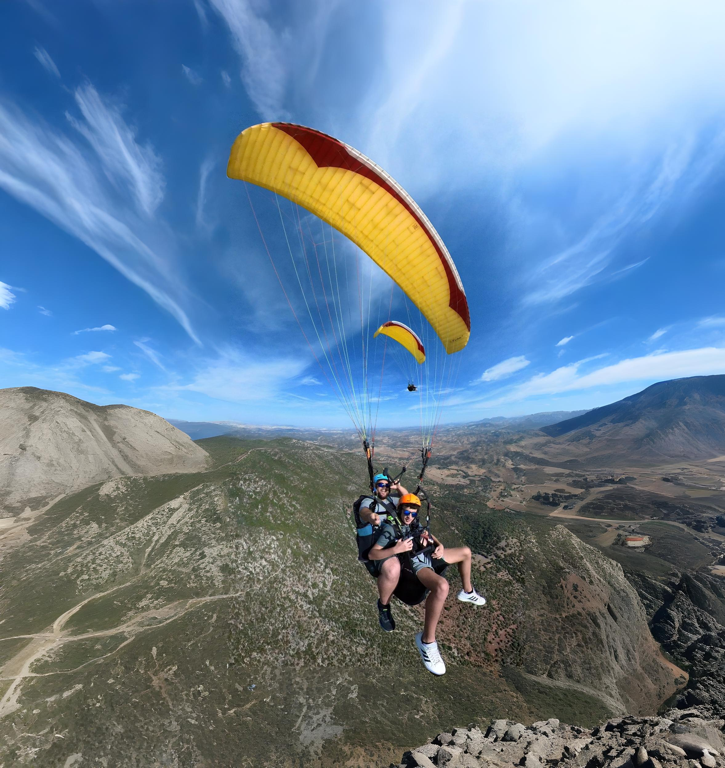Dos personas volando en parapente biplaza con un ala amarilla sobre un paisaje montañoso, bajo un cielo azul con nubes finas y dispersas.