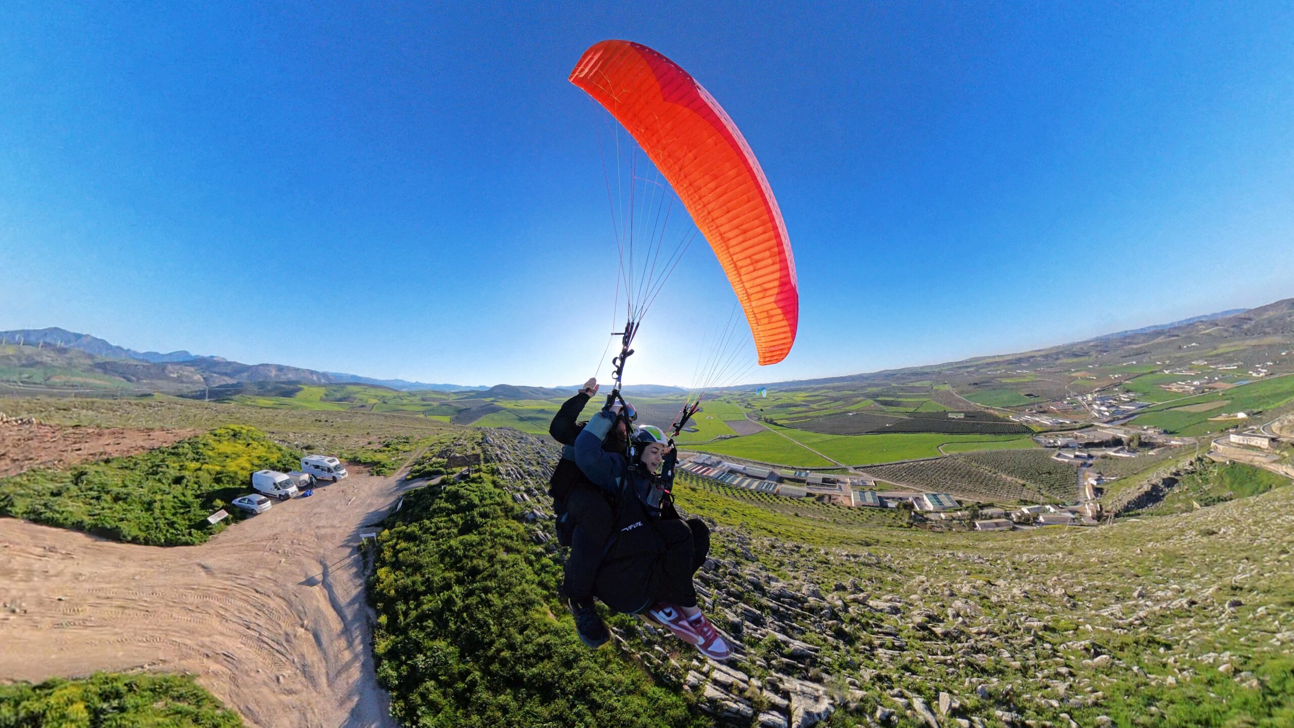 Persona despegando en parapente con una vela roja sobre un paisaje rural de colinas verdes, bajo un cielo completamente despejado.