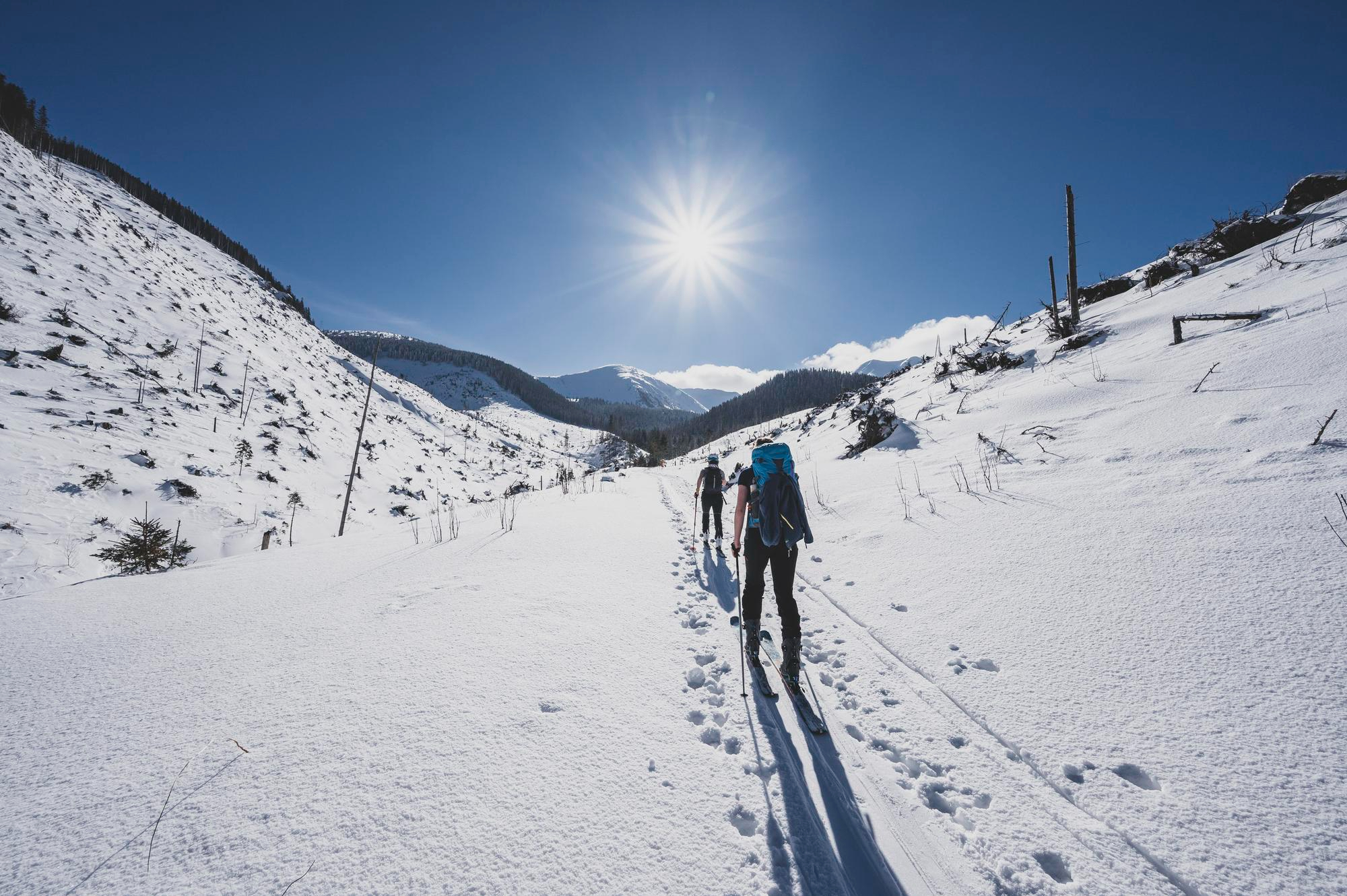 Pareja de excursionistas avanzando por un sendero cubierto de nieve en la montaña, con mochilas y bastones, rodeados de colinas nevadas y cielo despejado.