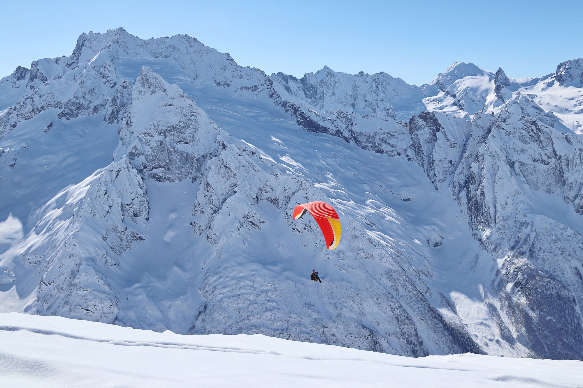 Un parapente de color rojo y amarillo volando frente a una inmensa cadena de montañas totalmente cubiertas de nieve bajo un cielo despejado.