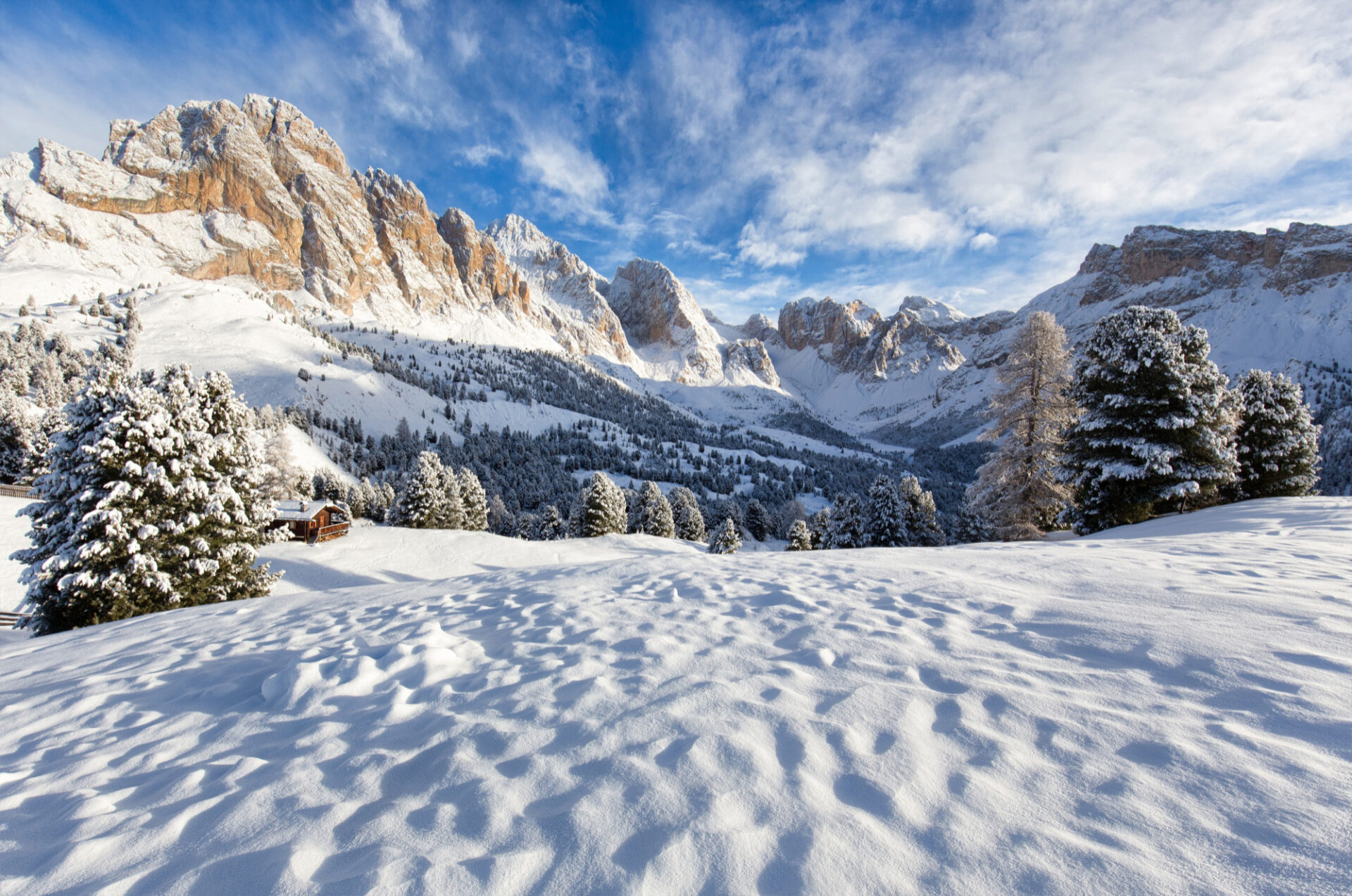 Paisaje invernal de alta montaña con picos rocosos nevados, un bosque de pinos cubiertos de nieve y una pequeña cabaña de madera bajo un cielo azul con nubes blancas.