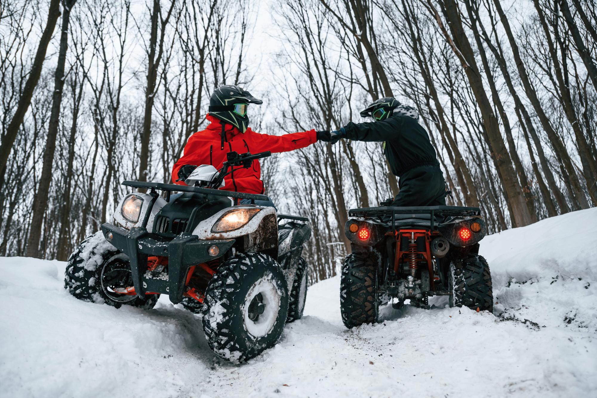 Dos personas con cascos y equipamiento de invierno chocando los puños sobre sus quads en un sendero nevado rodeado de árboles.