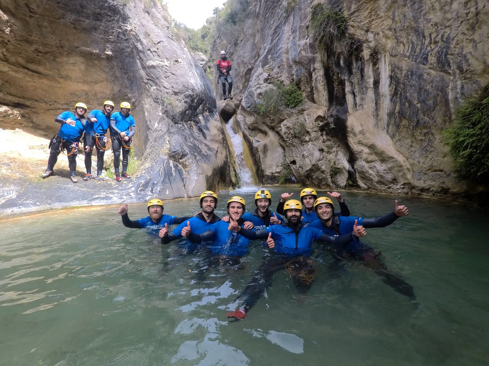 Un grupo de personas sonrientes practicando barranquismo en un cañón natural. Visten neoprenos azules y cascos amarillos, algunos están sumergidos en el agua turquesa y otros posan sobre las rocas. Al fondo se ve una pequeña cascada entre paredes de roca escarpadas.