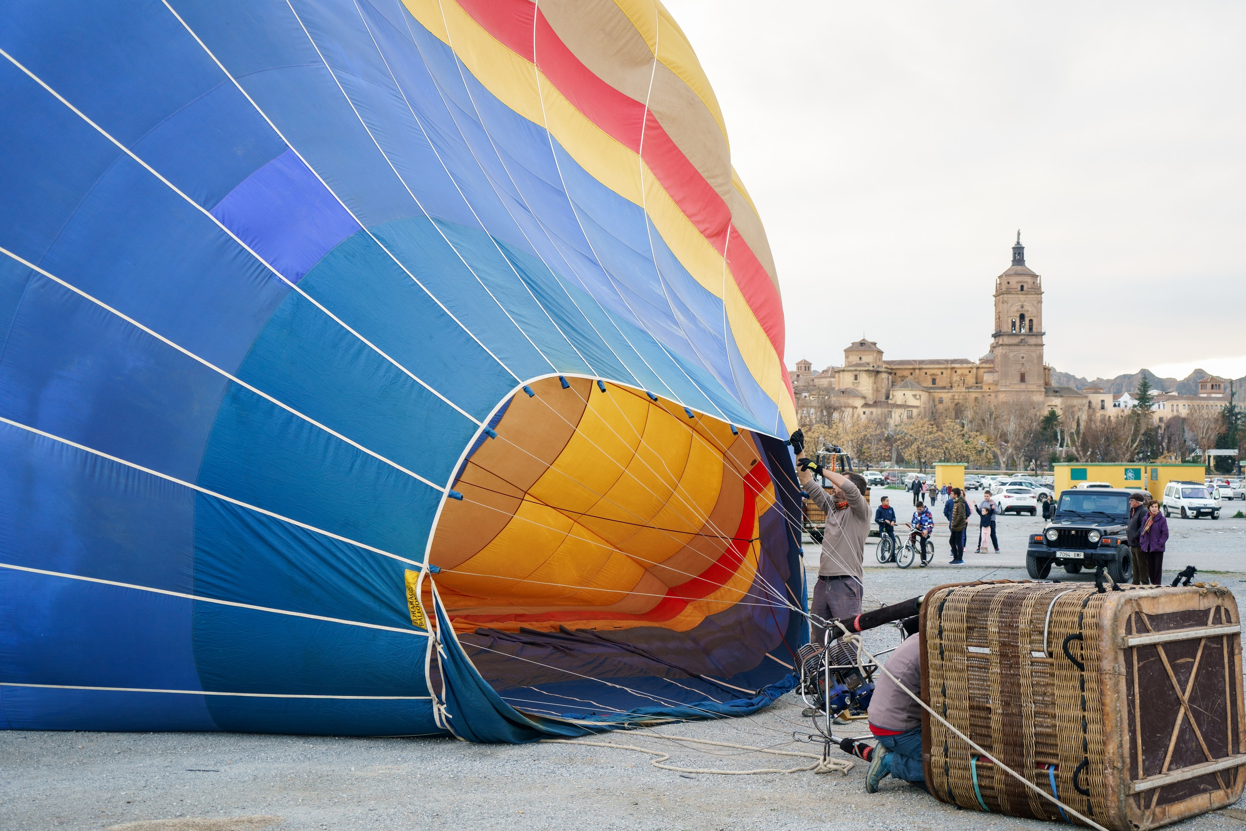 Un primer plano de un gran globo aerostático de colores azul, amarillo y rojo siendo inflado en un terreno abierto en Guadix, Granada. Un operario sostiene la apertura del globo mientras otro trabaja cerca de la cesta de mimbre volcada. Al fondo, se aprecia la silueta histórica de la Catedral de Guadix bajo un cielo nublado.