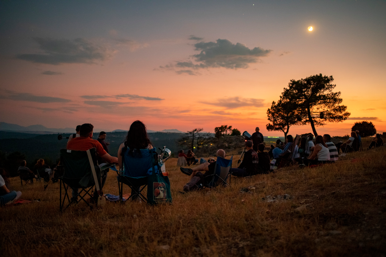 Un grupo de personas sentadas en una ladera, algunas en sillas de camping, observando el atardecer y la salida de la luna en un entorno rural despejado.