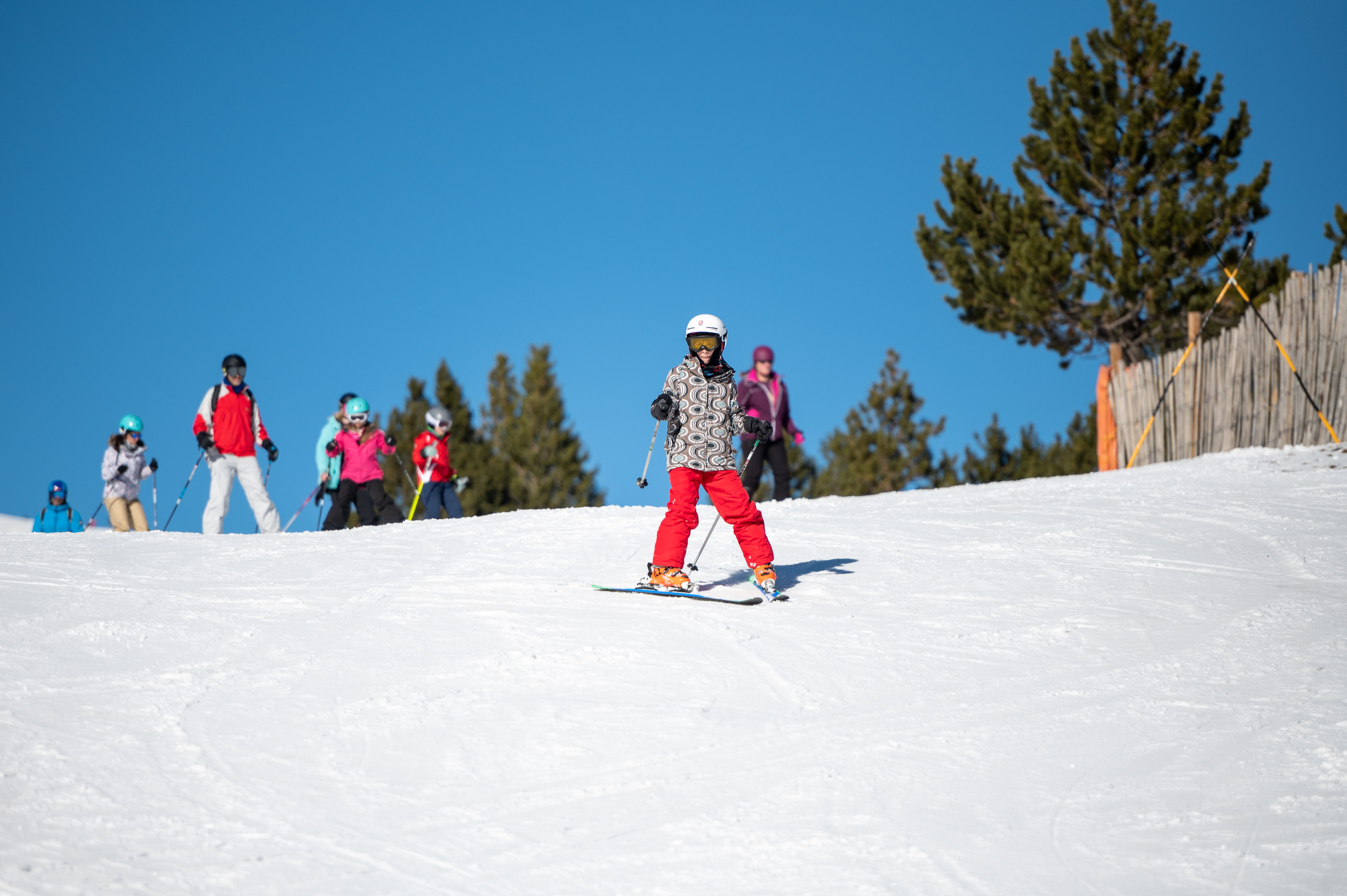 Persona principiante practicando esquí en una pista soleada de España durante una clase de esquí, con otros esquiadores y un monitor al fondo.