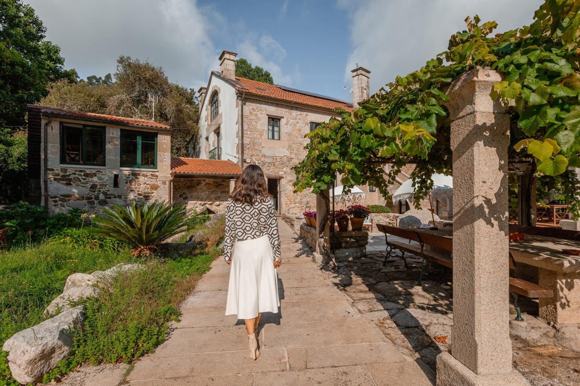 Vista posterior de una mujer caminando por un sendero empedrado hacia una casa rural de piedra tradicional, con un cenador de parras a la derecha y vegetación frondosa alrededor.