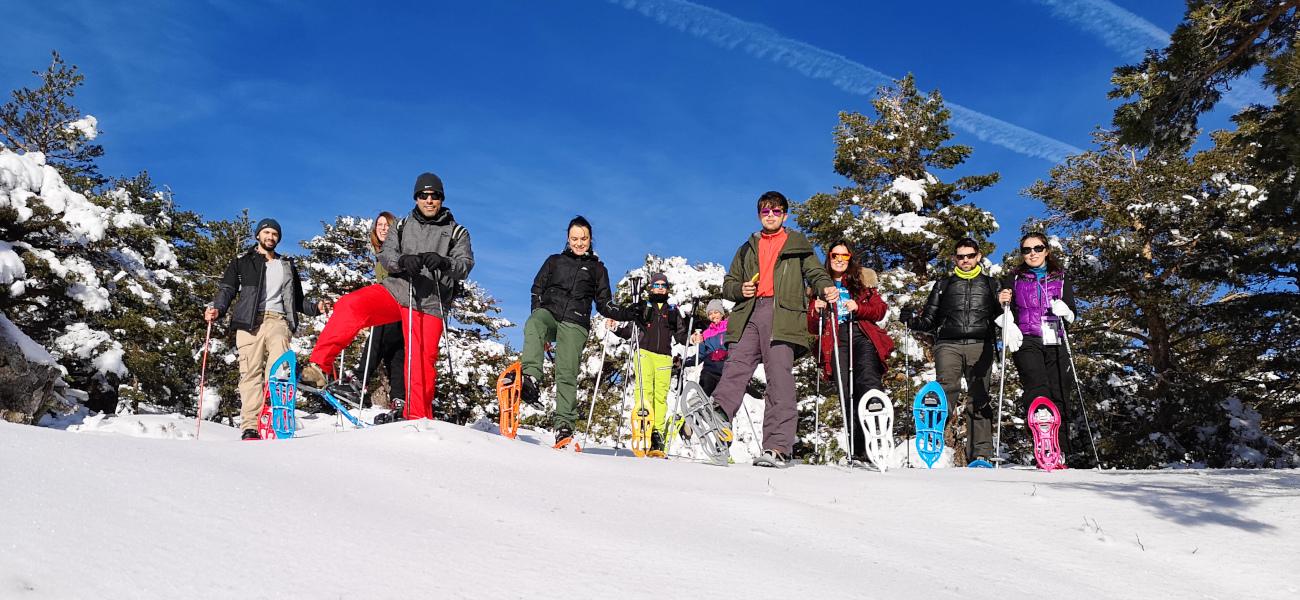 Un grupo de excursionistas posa con raquetas de nieve en Sierra Nevada, Granada. Se encuentran en una ladera nevada rodeada de pinos bajo un cielo azul despejado, mostrando su equipo de invierno y bastones de trekking.