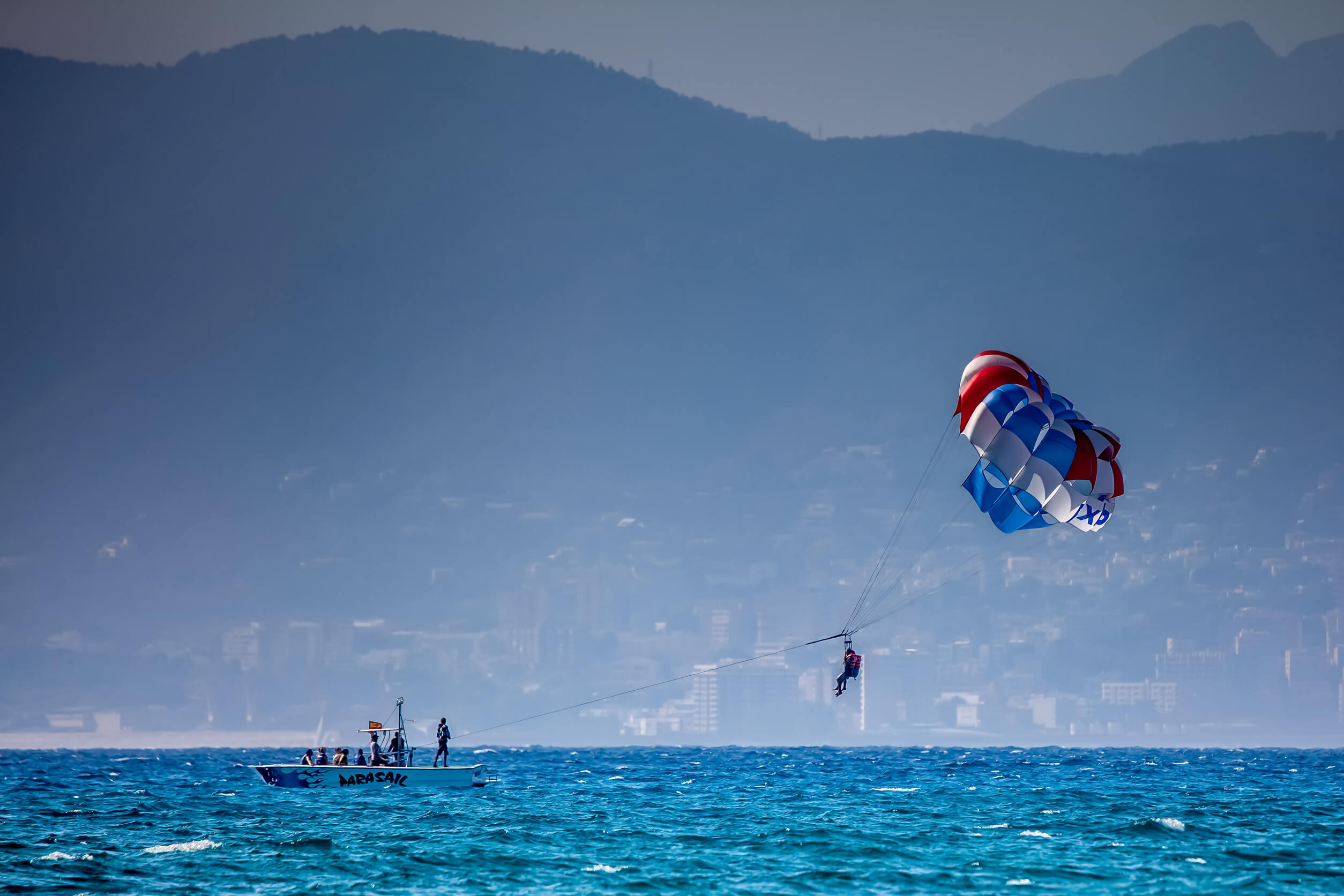 Una persona practicando paracaidismo (parasailing) sobre el mar, remolcada por una lancha con montañas y una ciudad costera al fondo.