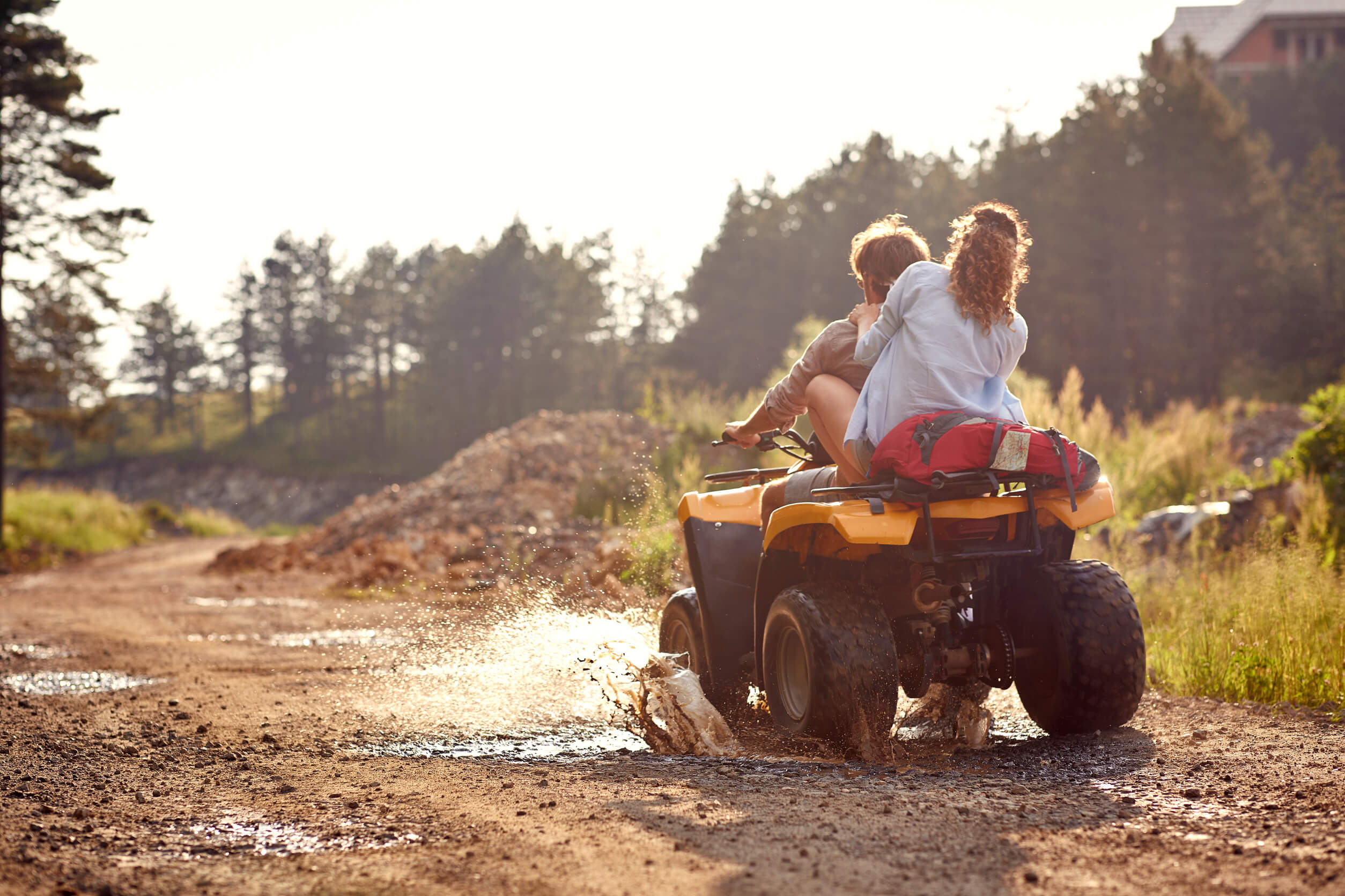 Una pareja en un quad amarillo cruzando un charco de agua y lodo en un camino forestal.