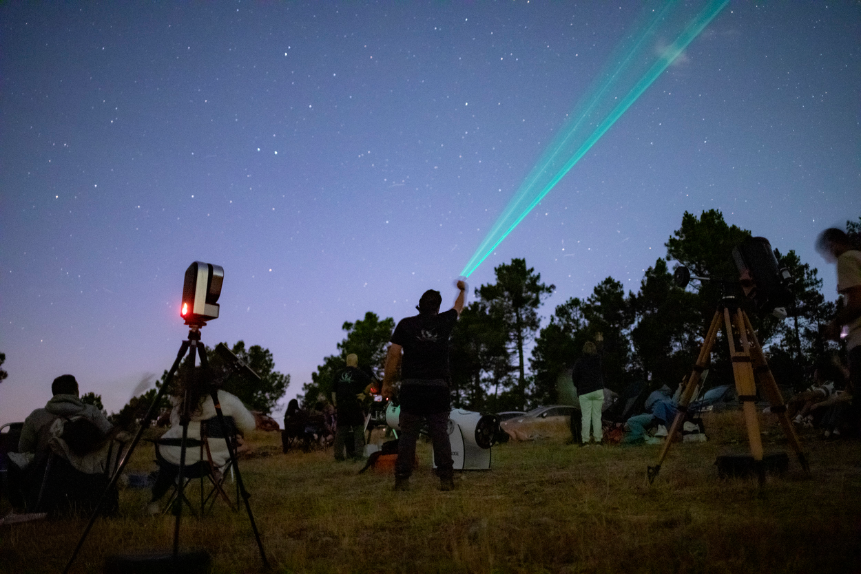 Un experto en astronomía utiliza un puntero láser verde para señalar constelaciones en el cielo nocturno ante un grupo de personas con telescopios en el campo