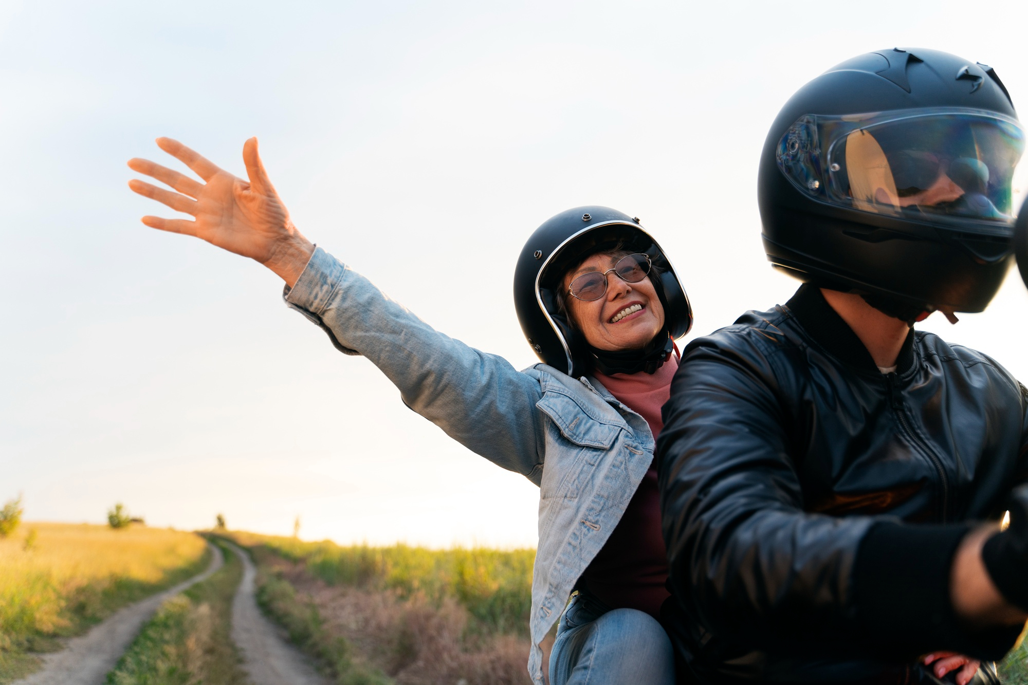 Pareja con cascos de moto, la pasajera sonriente saluda con la mano, conduciendo por un camino rural al atardecer