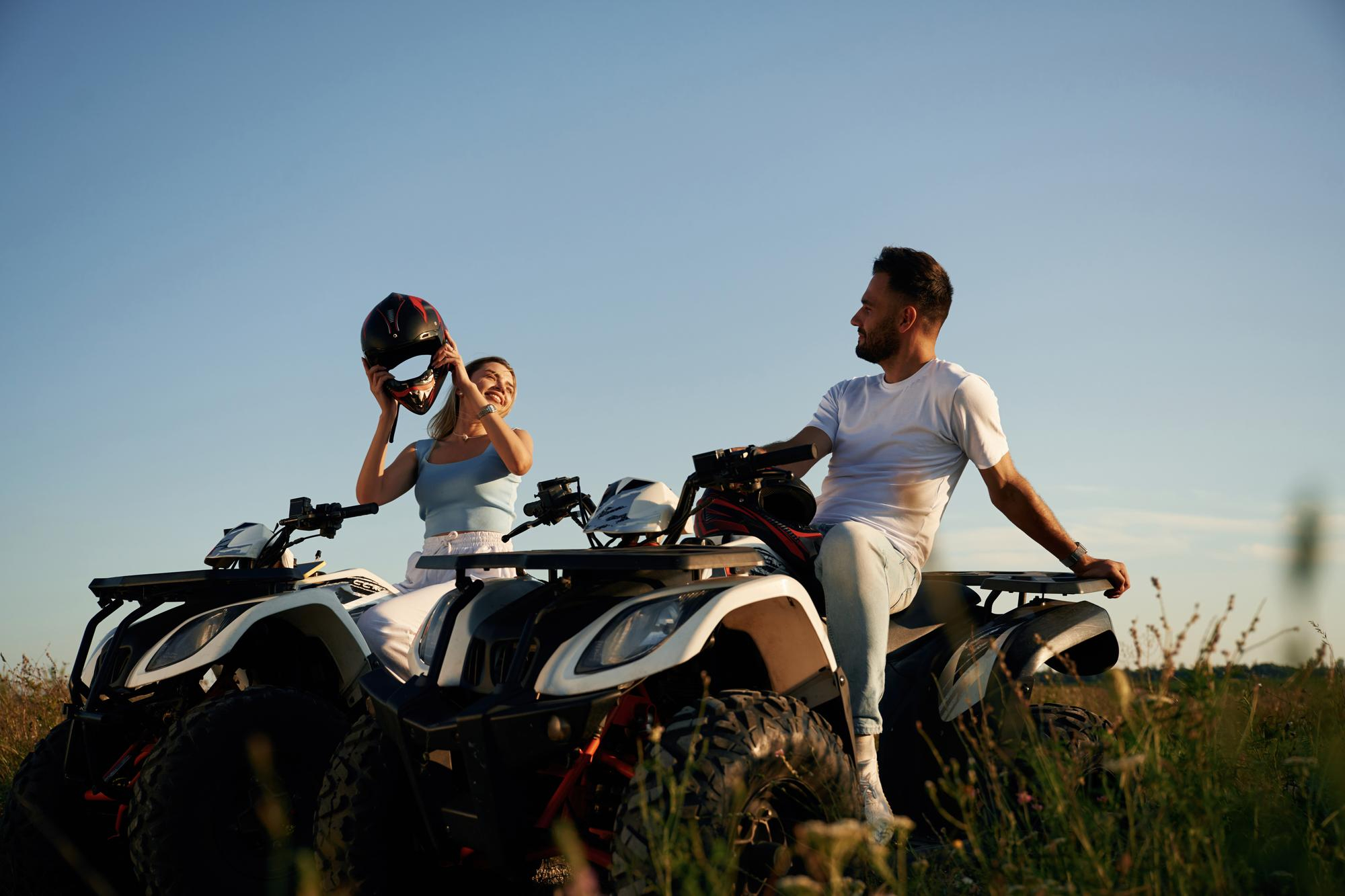 Pareja relajándose sobre dos quads en un campo al atardecer, con la mujer poniéndose un casco y el hombre mirándola