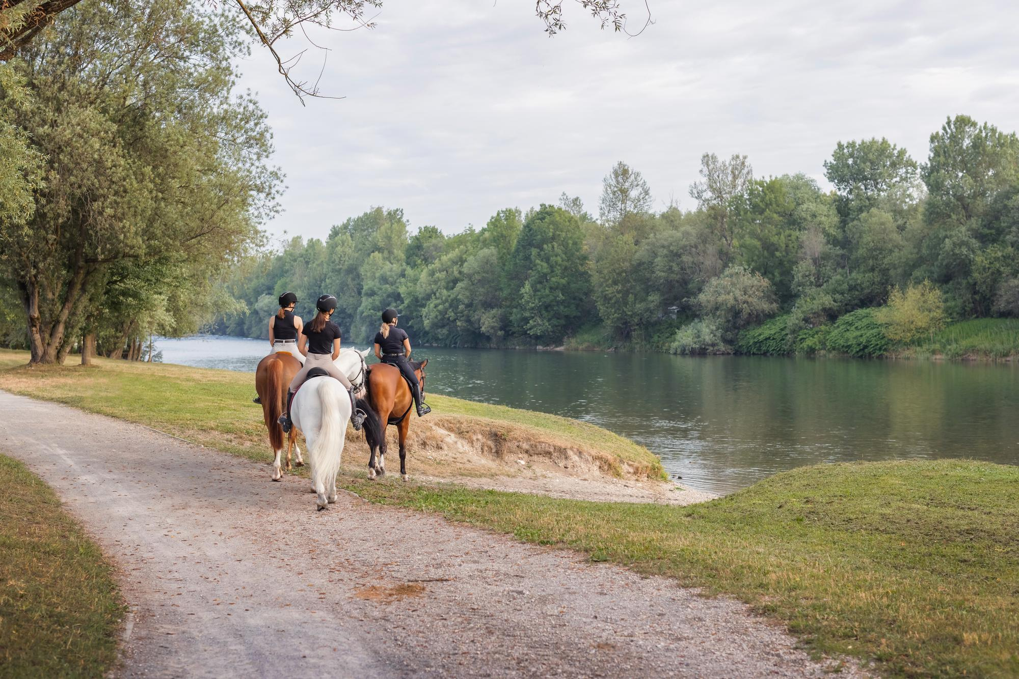 Tres personas montando a caballo por un sendero de tierra junto a un río tranquilo, rodeado de árboles y vegetación frondosa bajo un cielo despejado.