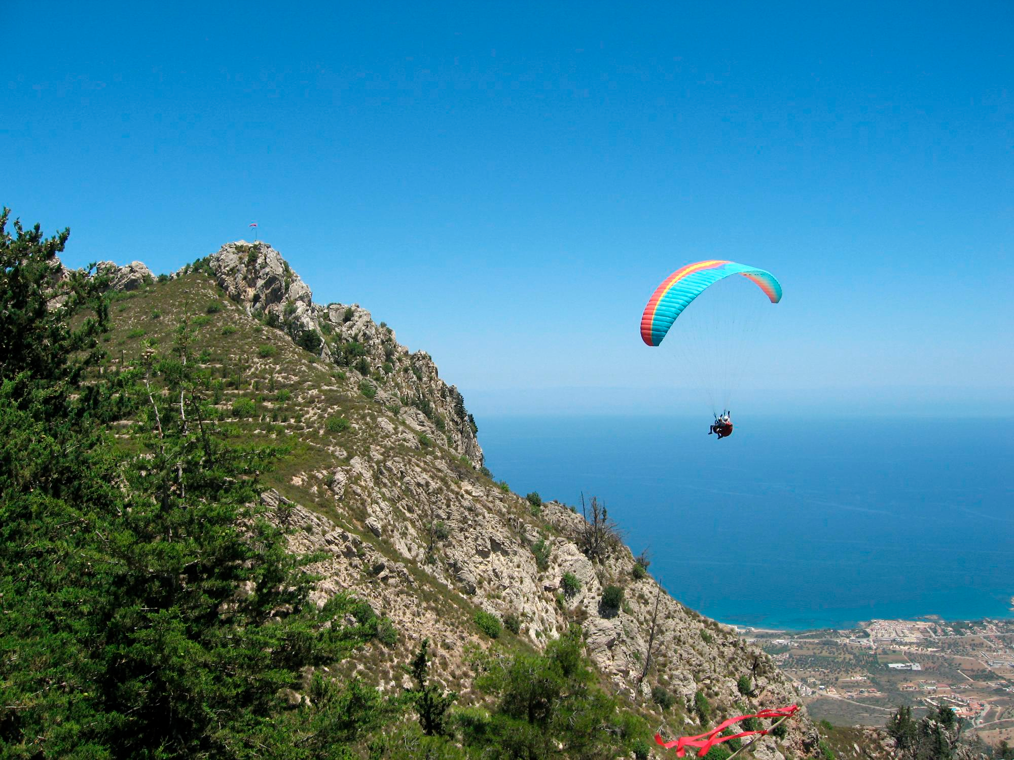 Vista de un parapente biplaza sobrevolando una montaña escarpada con el mar azul de fondo bajo un cielo despejado
