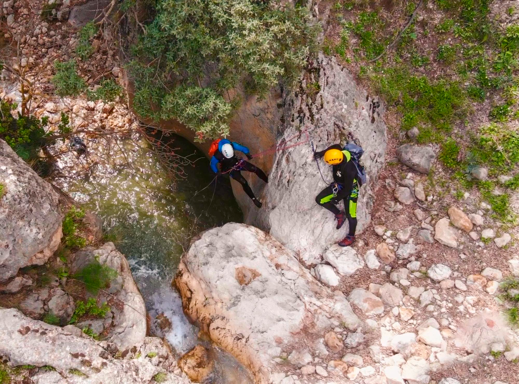 Dos personas con cascos y arneses descienden en rápel por una pared rocosa hacia una poza de agua cristalina en un barranco durante una actividad de barranquismo.
