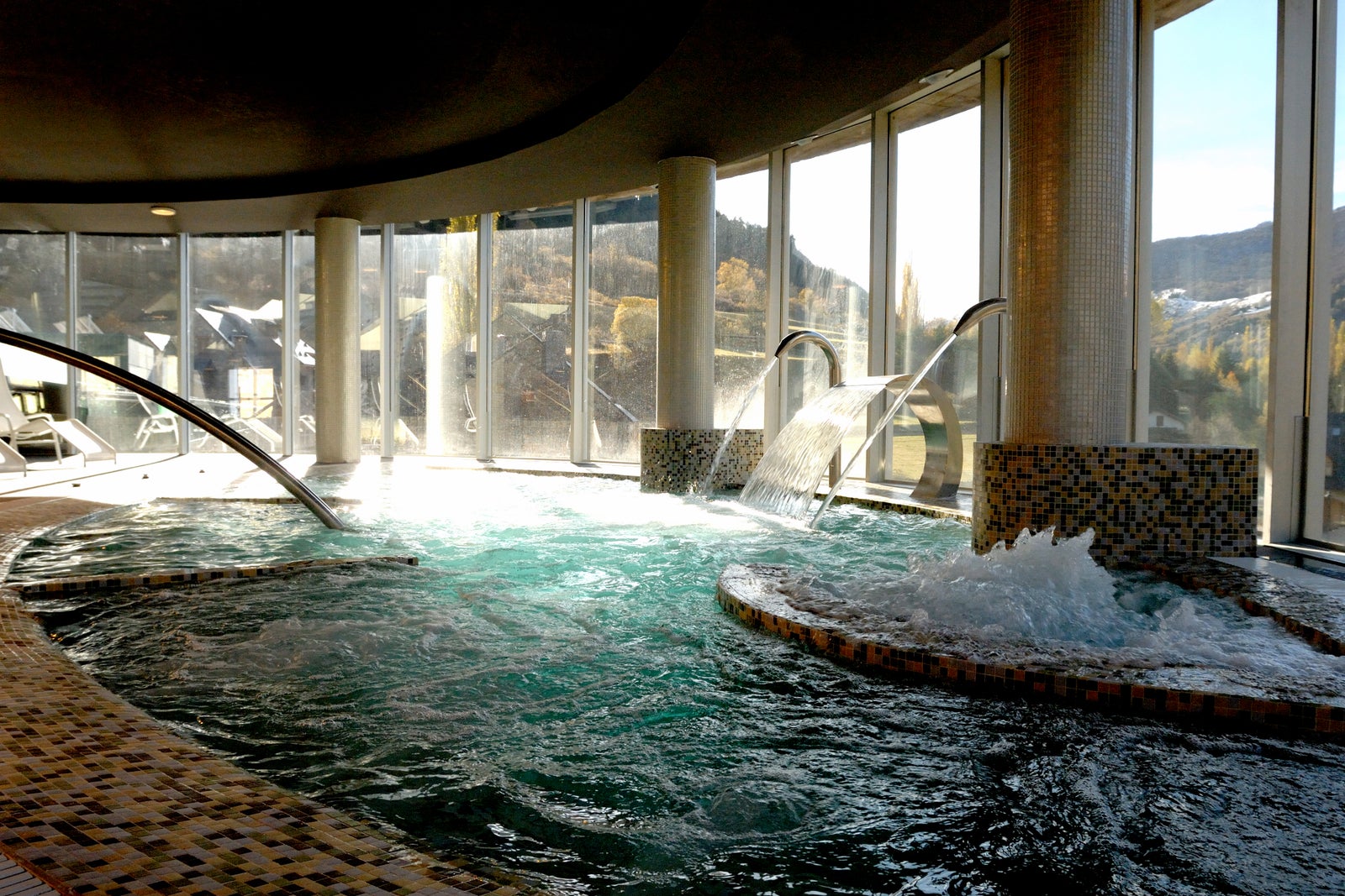 Circuito termal con piscinas de hidromasaje en un spa con grandes ventanales y vistas al exterior.