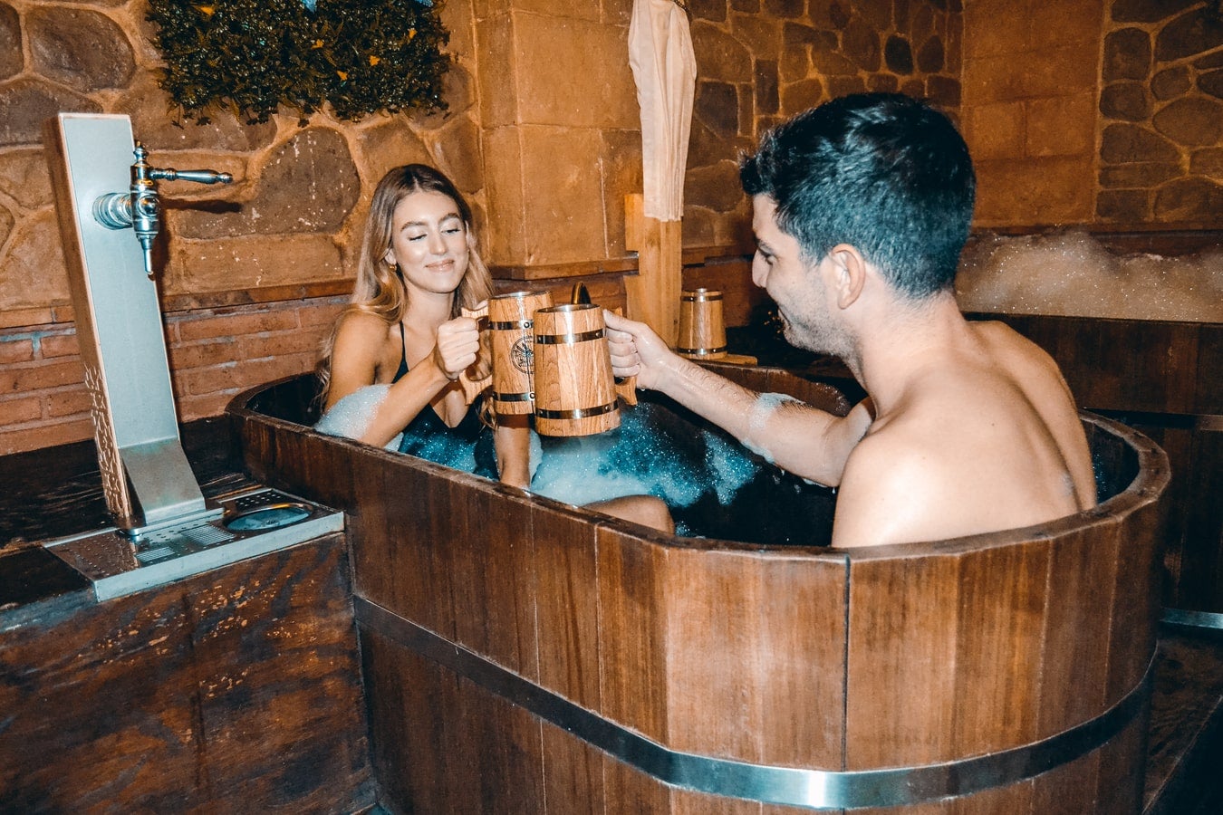 Una pareja se relaja en una tina de madera llena de espuma en un spa rústico, brindando con jarras de madera en un ambiente cálido con paredes de ladrillo