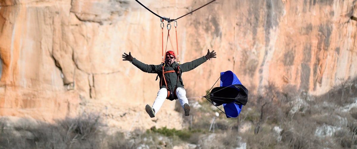 Hombre con los brazos abiertos descendiendo en tirolina con paracaídas de frenado sobre la hoz del río en Cuenca, con imponentes paredes de roca caliza al fondo