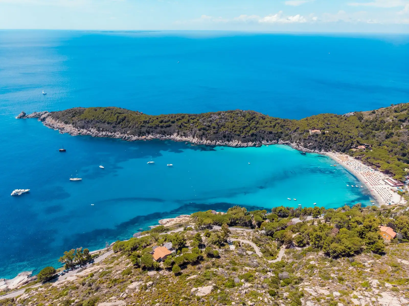 Vista aérea de una pintoresca bahía costera de aguas turquesas y una playa de arena blanca, rodeada de densos bosques de pinos bajo un cielo despejado.