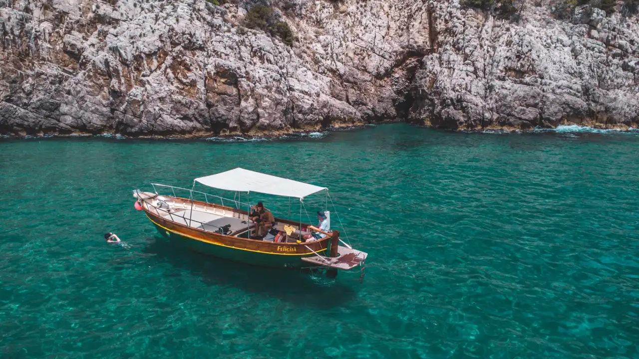 Pequeño barco de madera con toldo blanco navegando en aguas turquesas cristalinas frente a un acantilado rocoso, con personas a bordo y un nadador en el agua.