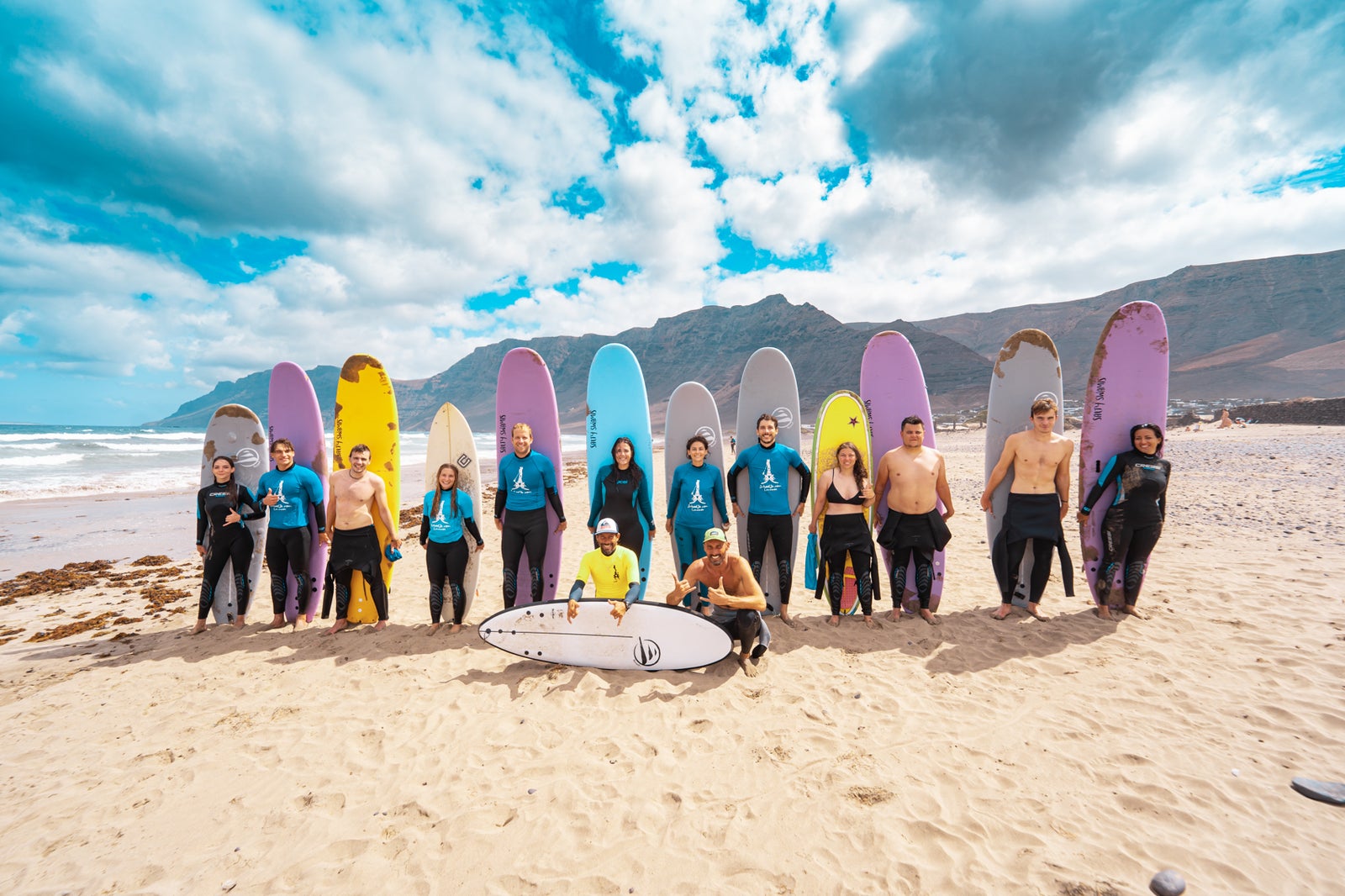 Un grupo de alumnos y monitores posando con sus tablas de surf de colores en una playa de arena clara, con montañas de fondo bajo un cielo parcialmente nublado