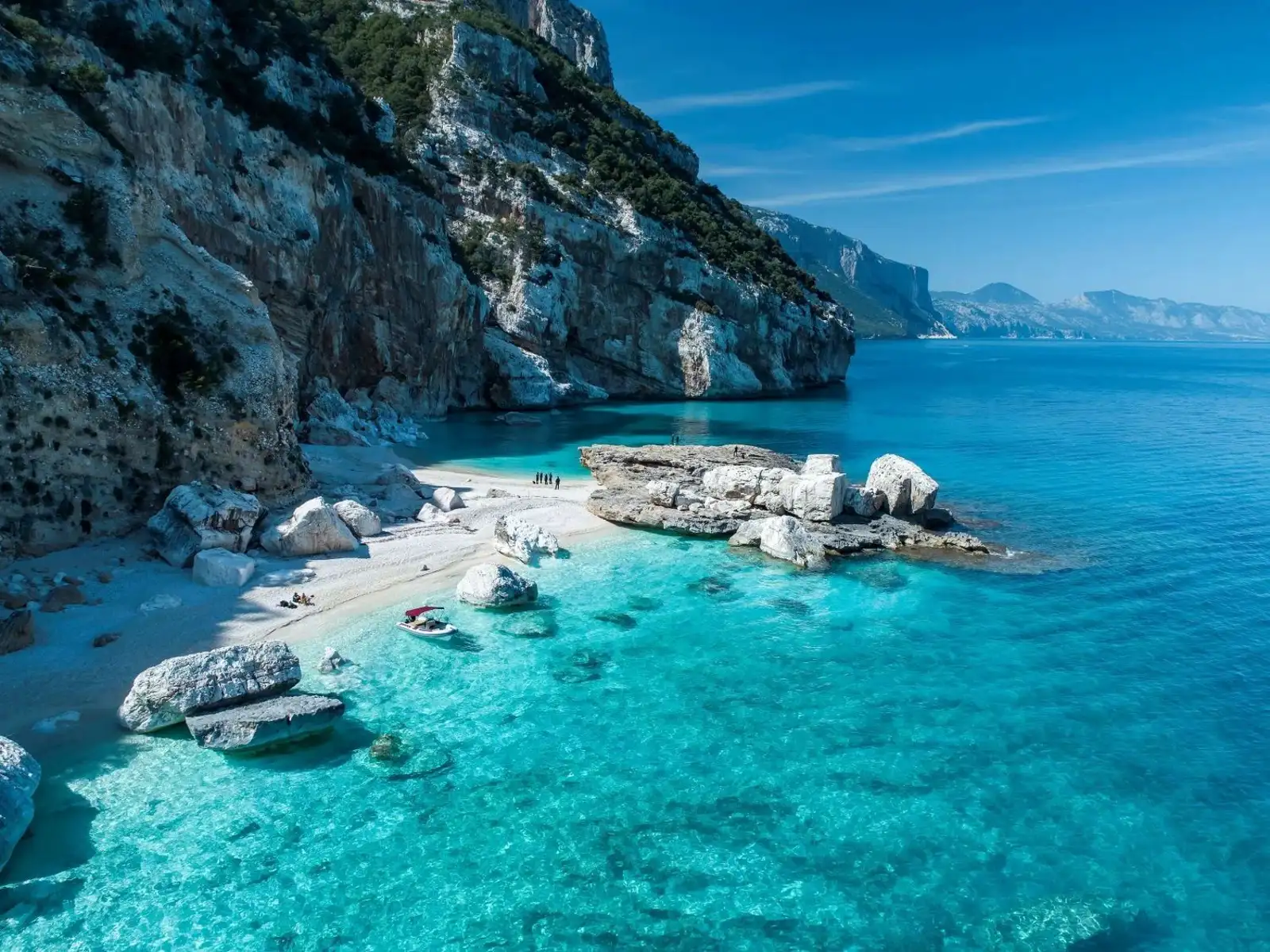Vista aérea de una espectacular playa de arena blanca y aguas turquesas cristalinas, rodeada de imponentes acantilados calizos con vegetación, rocas blancas dispersas en la orilla y una pequeña lancha fondeada en el mar.
