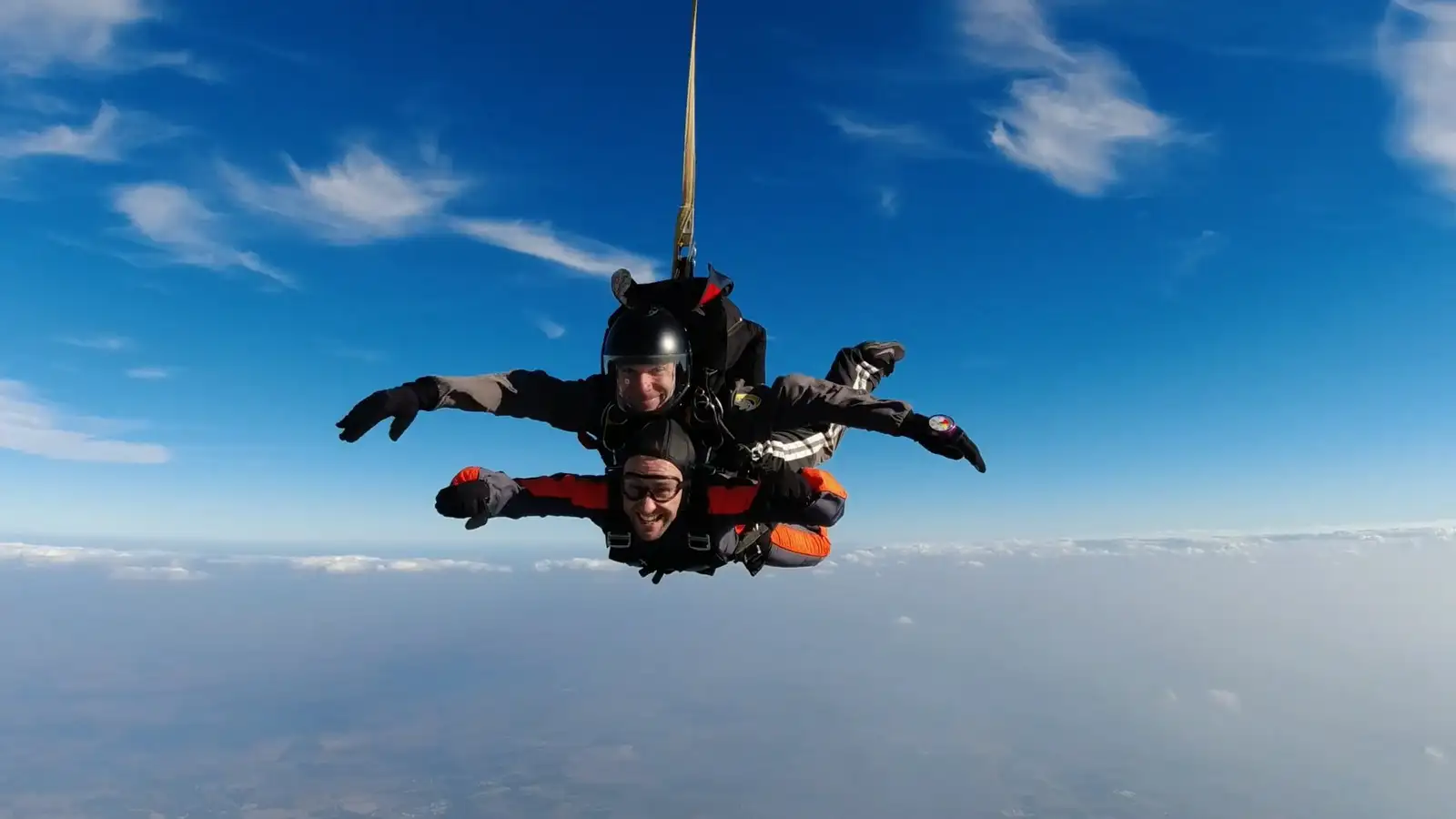 Instructor y alumno realizando un salto en tándem en paracaídas, con los brazos extendidos en posición de caída libre bajo un cielo azul despejado y sobre un manto de nubes.
