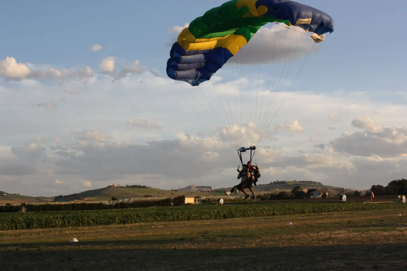 Paracaidista aterrizando suavemente con un paracaídas abierto de colores verde, amarillo y azul, aproximándose a una zona de césped con colinas al fondo bajo un cielo nublado