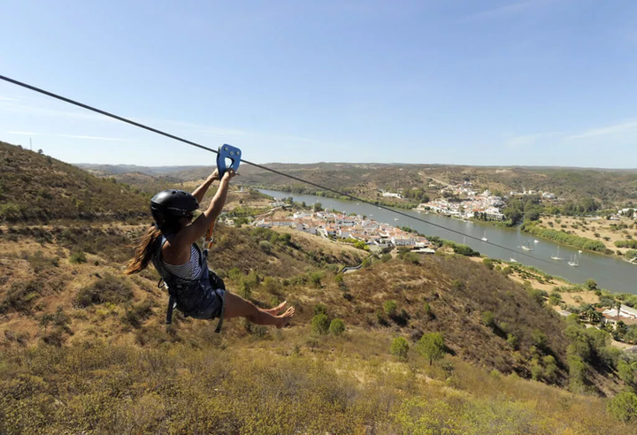 Mujer descendiendo en tirolina transfronteriza sobre el río Guadiana con vistas aéreas a un pueblo blanco ribereño y colinas del Algarve portugués bajo cielo despejado