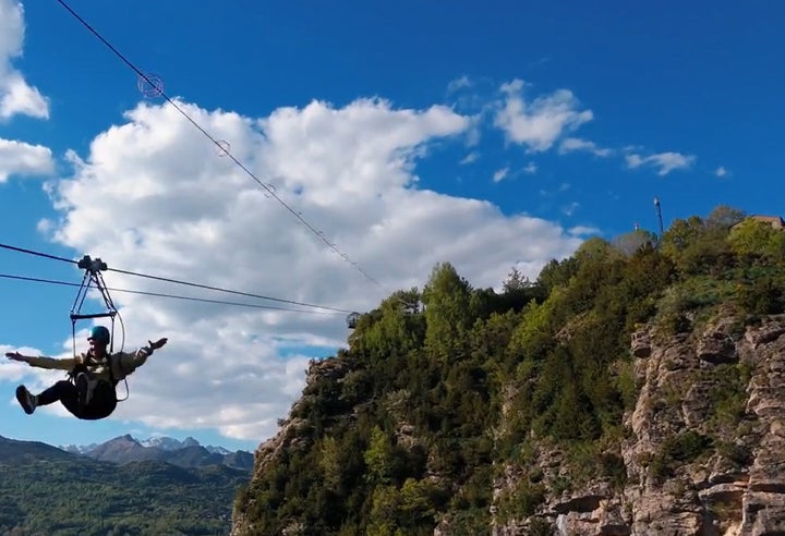 Persona deslizándose en tirolina sobre un barranco de montaña con los brazos abiertos, equipada con casco y arnés de seguridad, con paisaje de acantilados rocosos, vegetación verde y cumbres nevadas al fondo bajo un cielo azul con nubes
