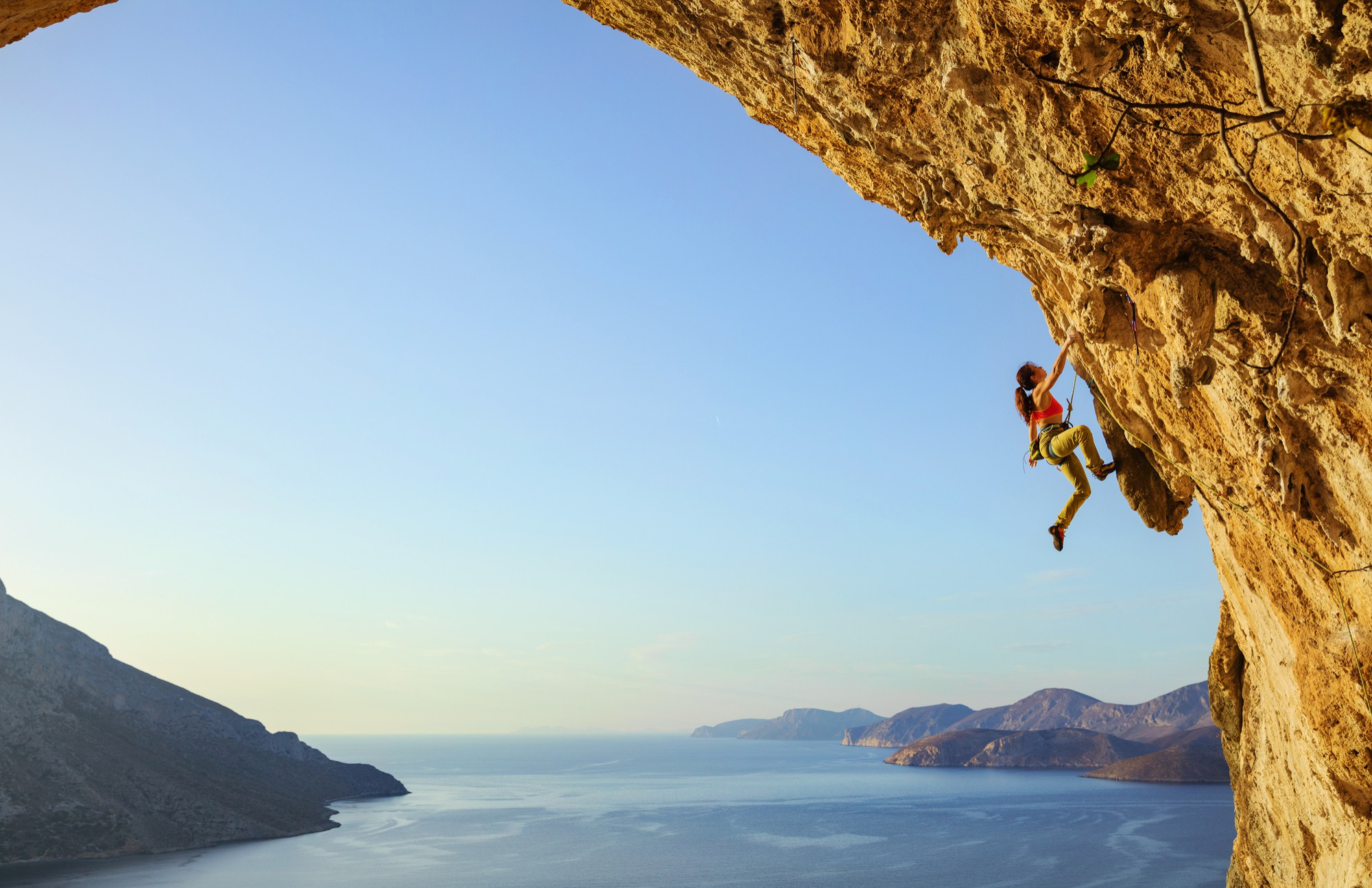 Una mujer escalando una pared de roca sobre un mar azul con montañas al fondo.