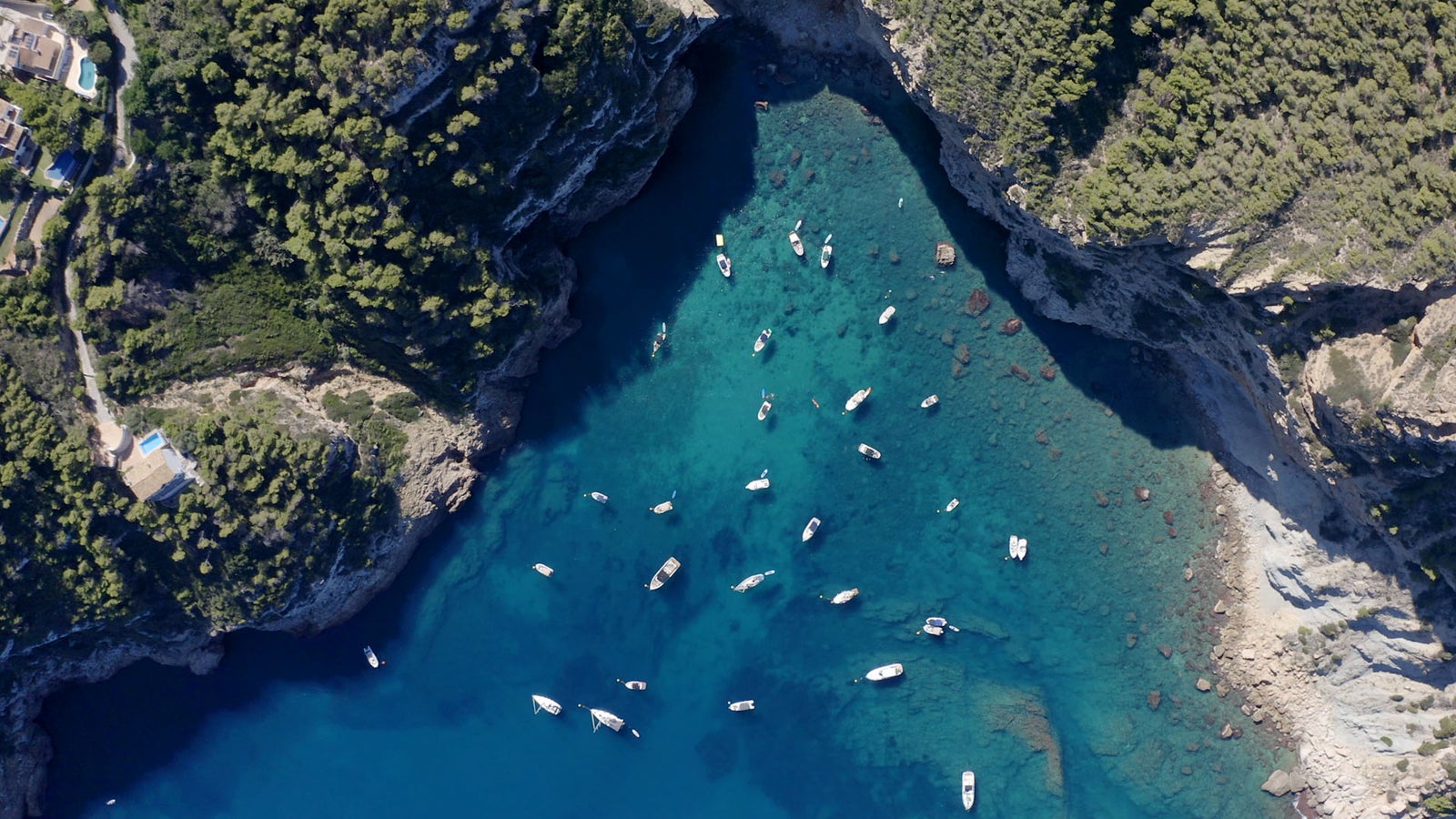 Vista aérea de una cala de aguas cristalinas turquesas con numerosos barcos y veleros fondeados, rodeada de acantilados y vegetación mediterránea.