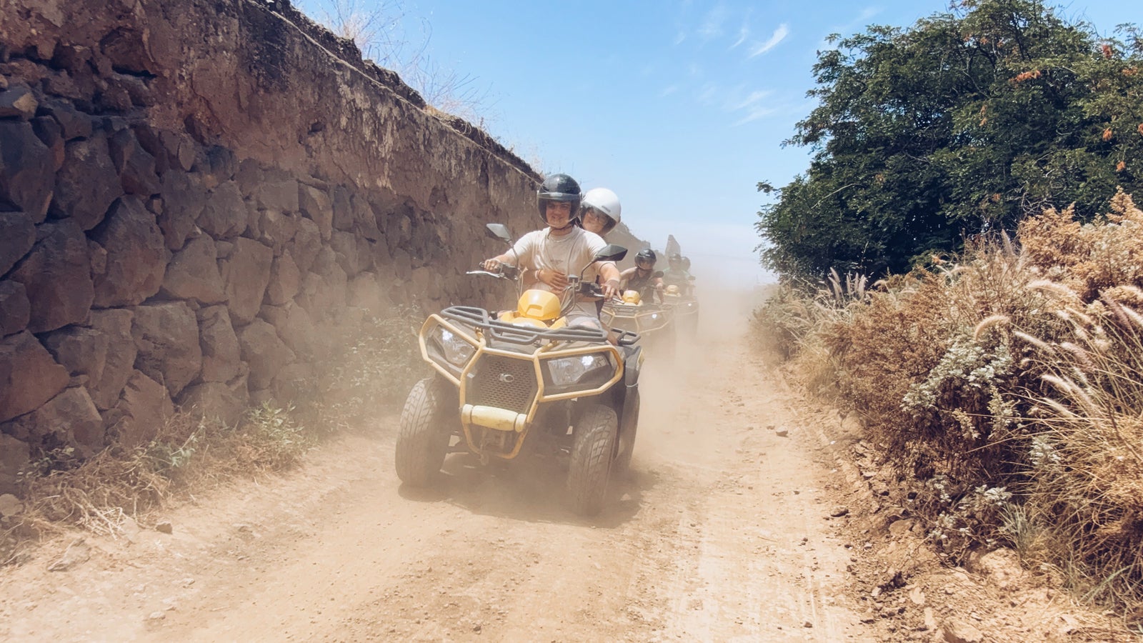 Un grupo de personas conduciendo quads por un camino de tierra polvoriento en Gran Canaria, con un paisaje árido y montañoso al fondo.