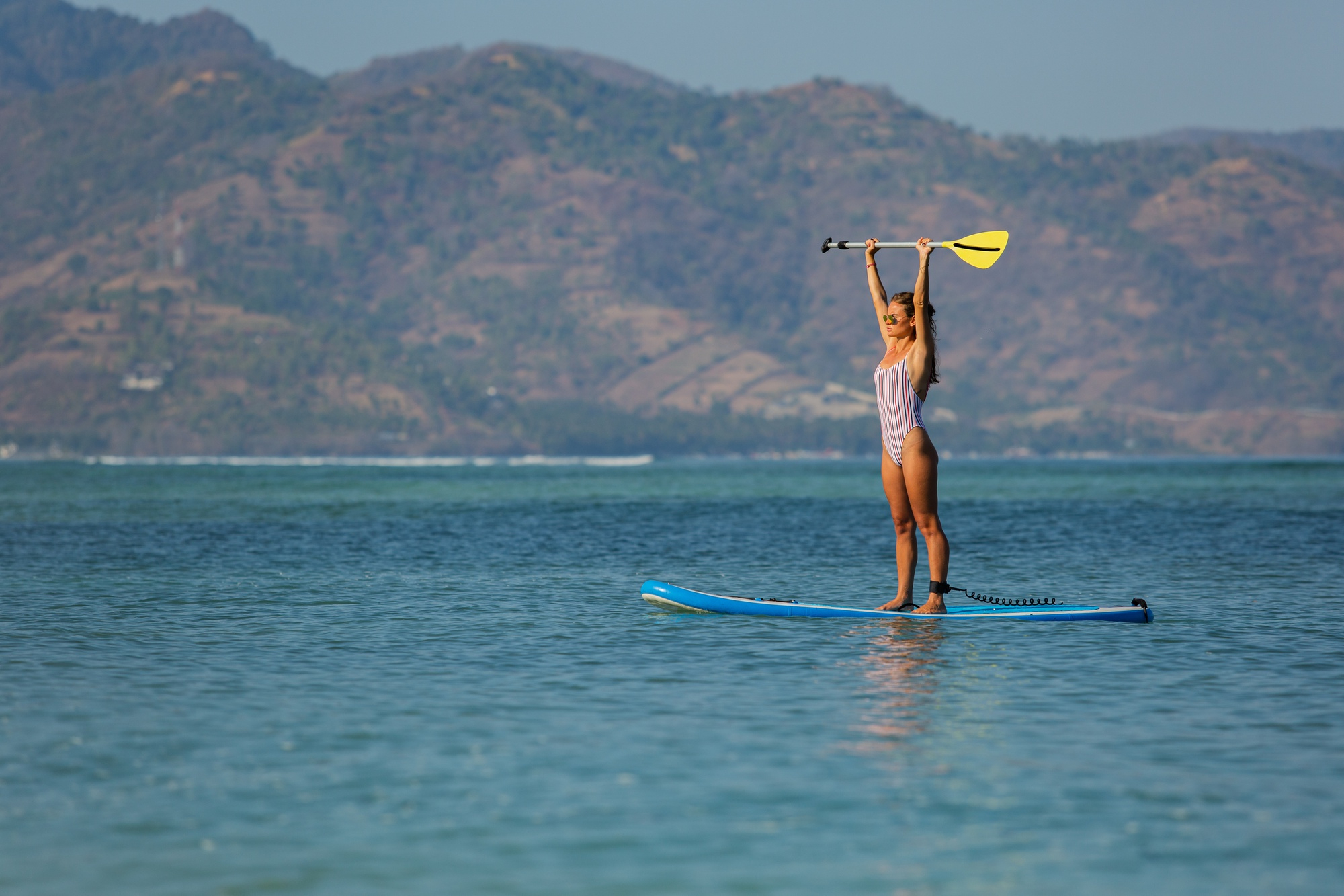Mujer en bañador rosa levantando el remo sobre una tabla de paddle surf con montañas de fondo