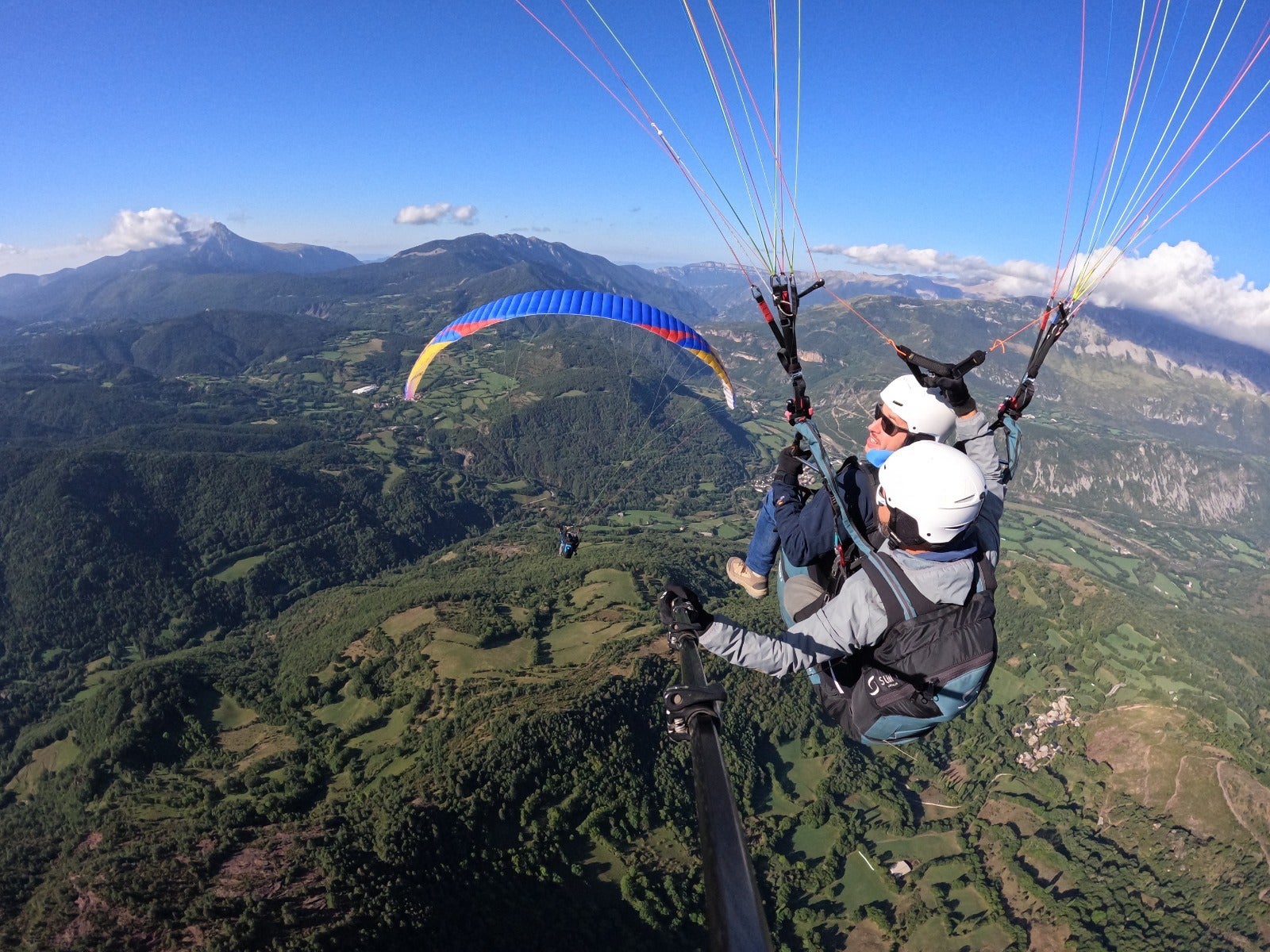 Dos personas vuelan en un parapente biplaza sobre un valle montañoso cubierto de bosques, bajo un cielo azul despejado.