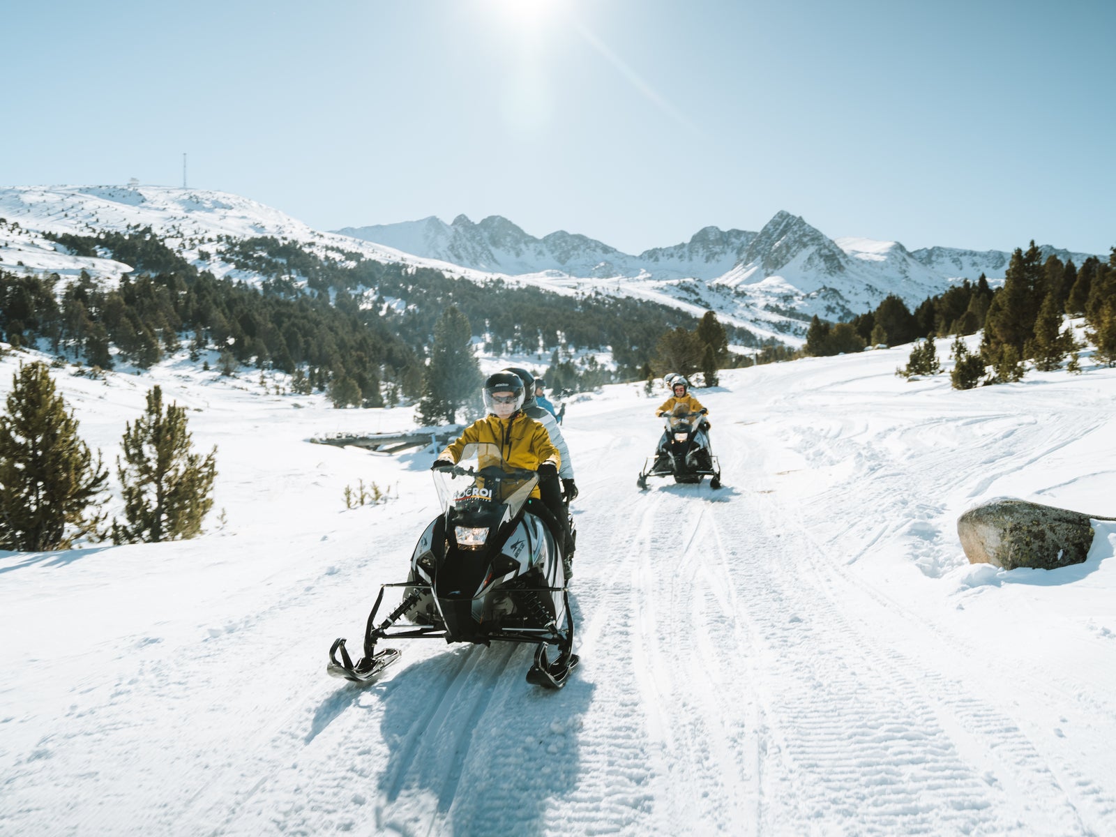Dos personas conducen motos de nieve por un camino nevado entre pinos, bajo un cielo soleado con montañas al fondo.