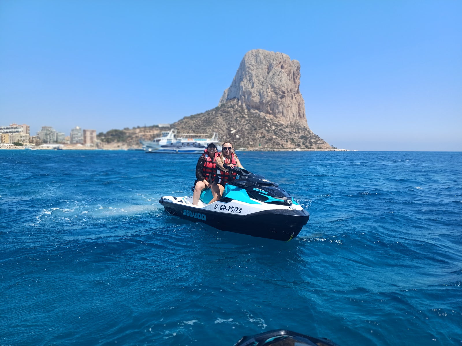 Pareja disfrutando de un paseo en moto de agua por el mar azul, con el Peñón de Ifach y la costa de Calpe al fondo bajo un cielo despejado.